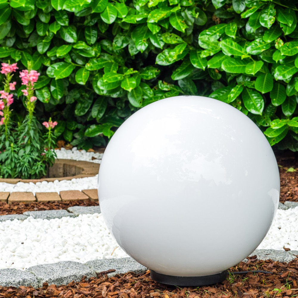 Eine weiße Kugel-Gartenleuchte steht im Vordergrund vor einem grünen Busch.