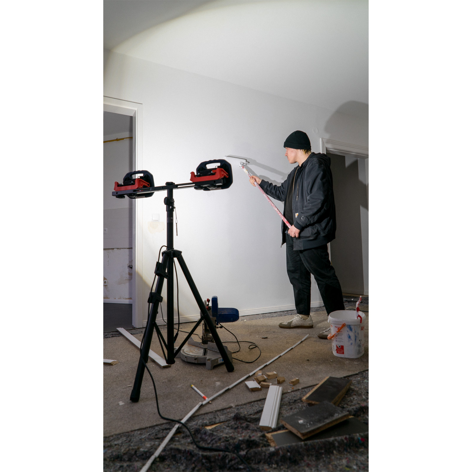 Ein Mann mit Mütze malt eine Wand mit einer Farbrolle, Werkzeuge und Farbeimer liegen herum.