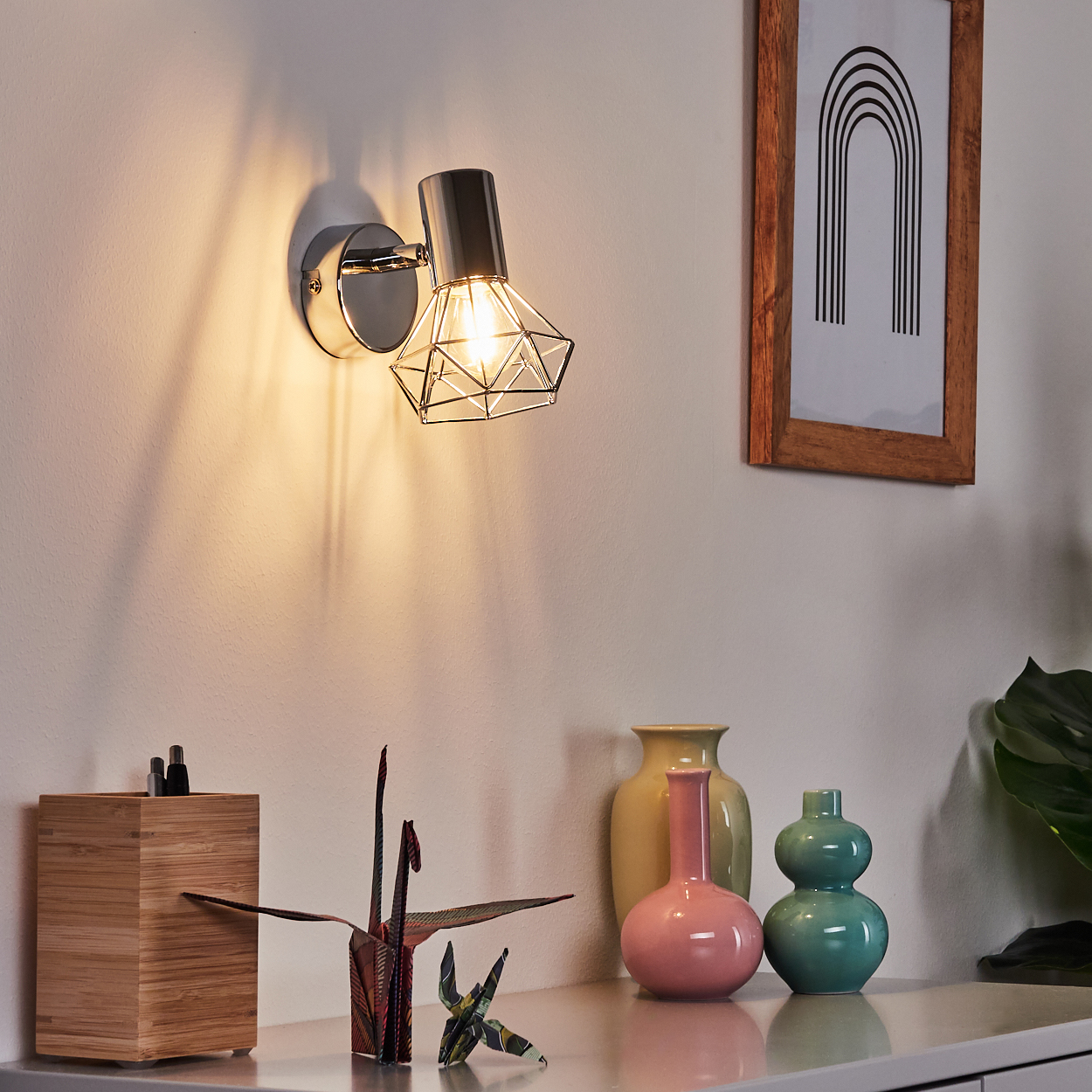 Eine Wandleuchte mit einem geometrischen Lampenschirm beleuchtet eine Wand. Dekorationen auf einem Regal.