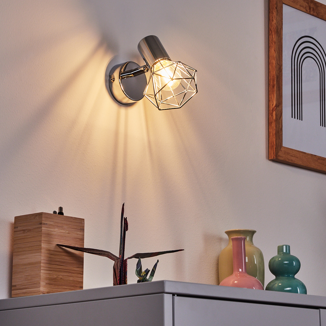 Ein moderner Wandspot mit Drahtkäfig, der auf einem Regal mit Vasen und Origami leuchtet.