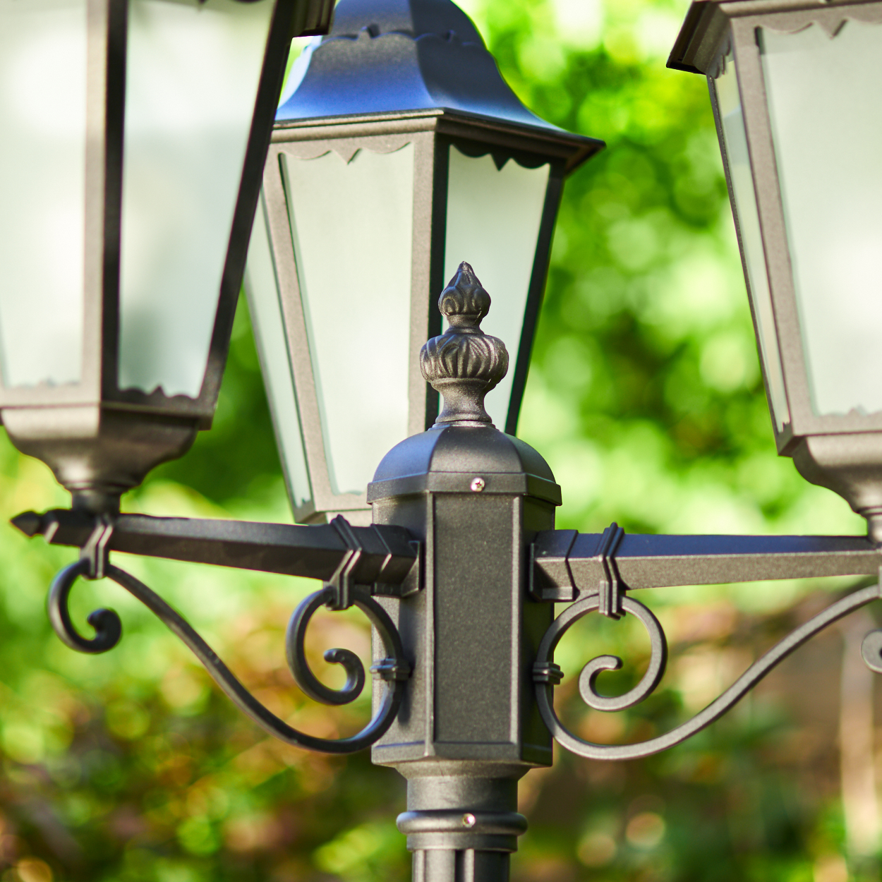 Ein schwarzer Laternenpfahl mit drei Lampen vor grünem, unscharfem Hintergrund.