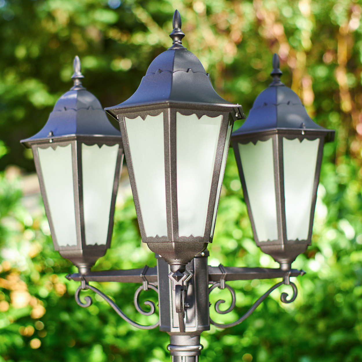Eine Laterne mit drei Lampen steht im Freien, umgeben von grünen Pflanzen.