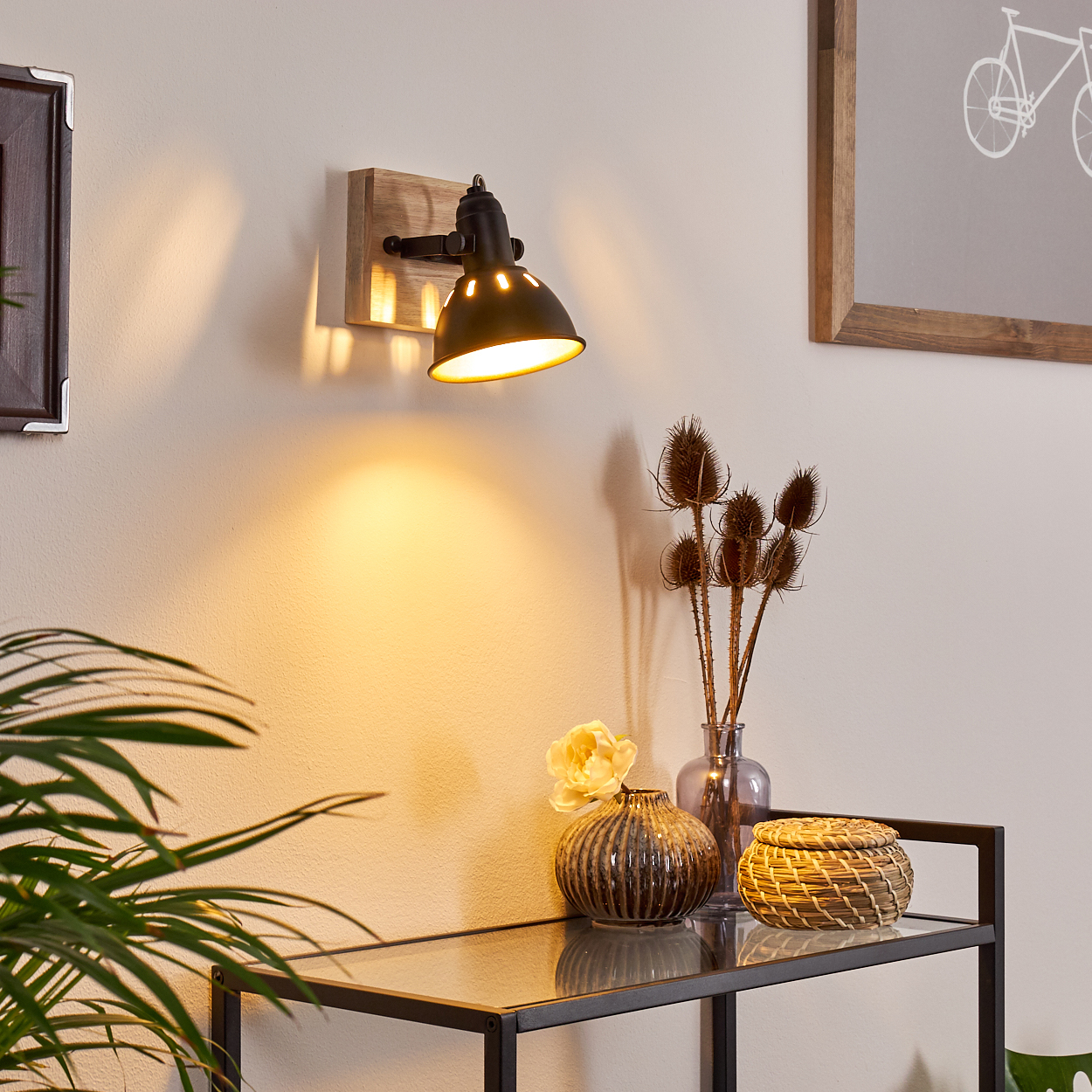 Ein schwarzer Wandspot mit Holzsockel beleuchtet eine Wand. Daneben stehen Vasen mit Trockenblumen.