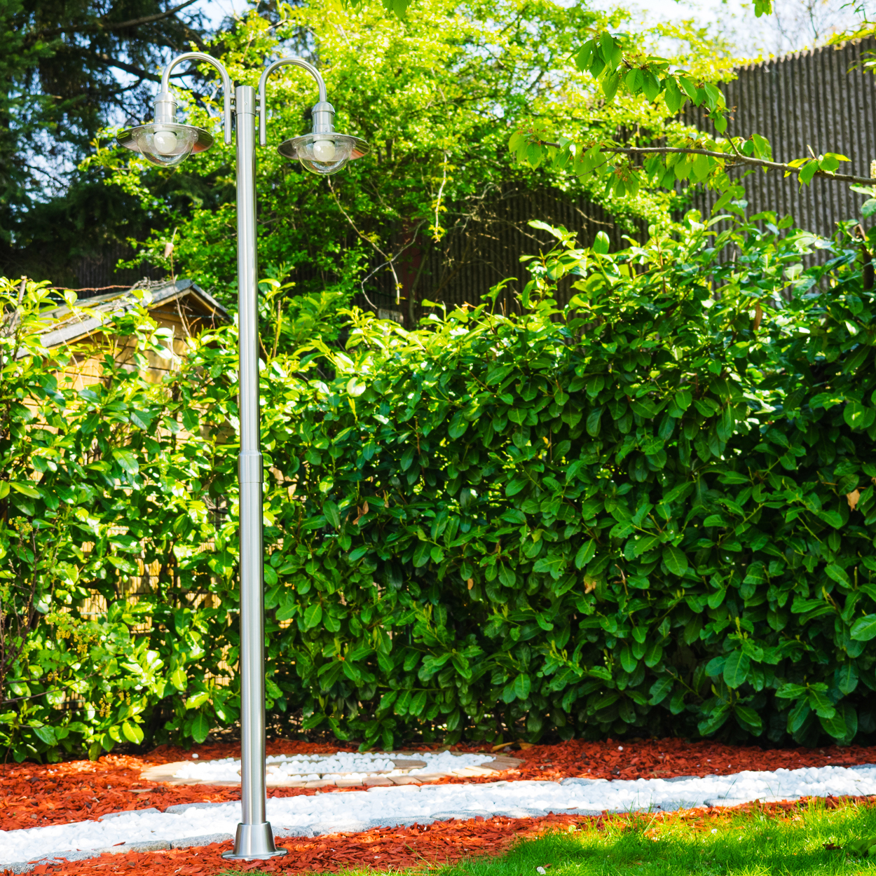 Zweiflammige Gartenlampe aus Metall mit Glaskuppeln, umgeben von grünen Hecken und Rindenmulch.