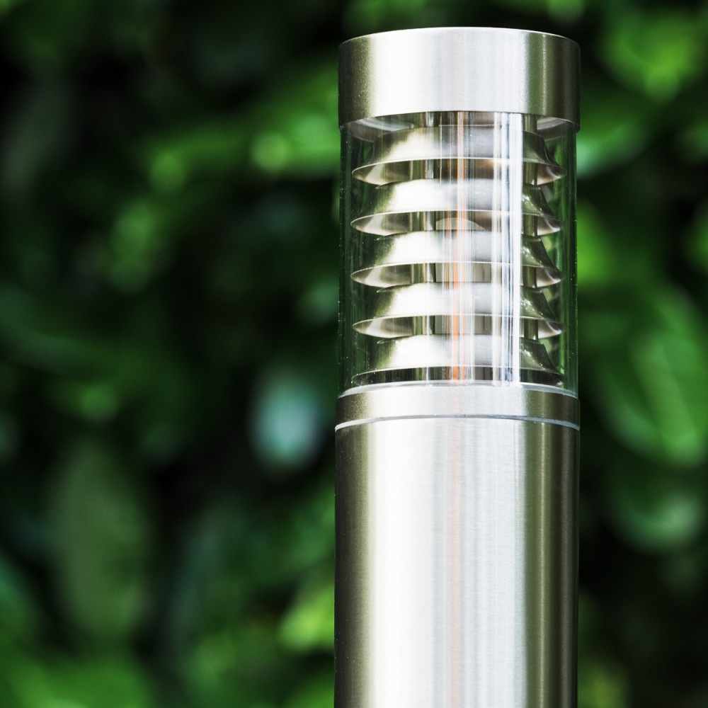 Moderne Edelstahl-Gartenleuchte mit Glaszylinder vor unscharfem grünen Hintergrund.