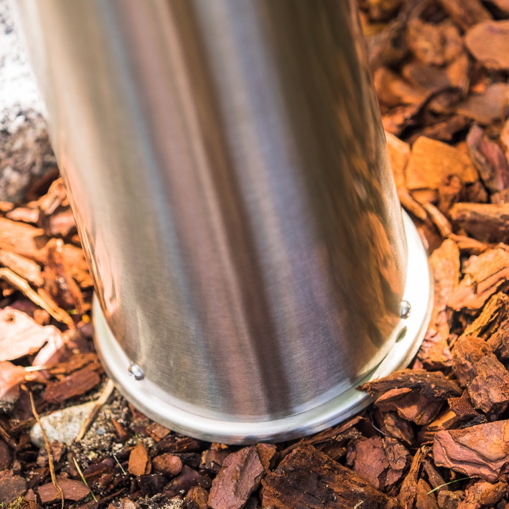 Ein polierter Metallpfosten steht auf Rindenmulch. Drei Nieten sind sichtbar.