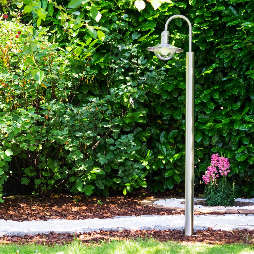 Ein Garten mit einem Metallpfosten und einer Lampe, grünen Büschen und rosa Blumen.
