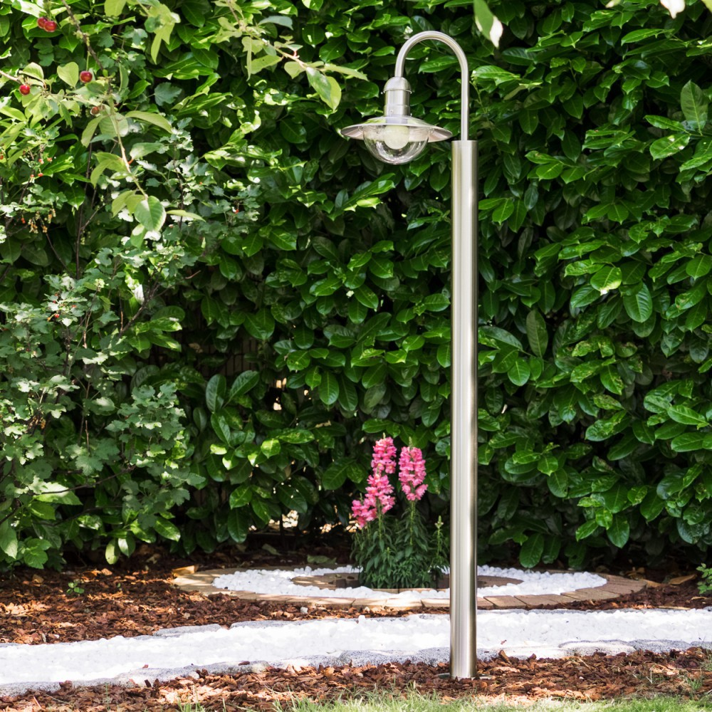 Ein moderner Edelstahl-Gartenlichtpfosten steht vor einer dichten grünen Hecke mit Blumen.