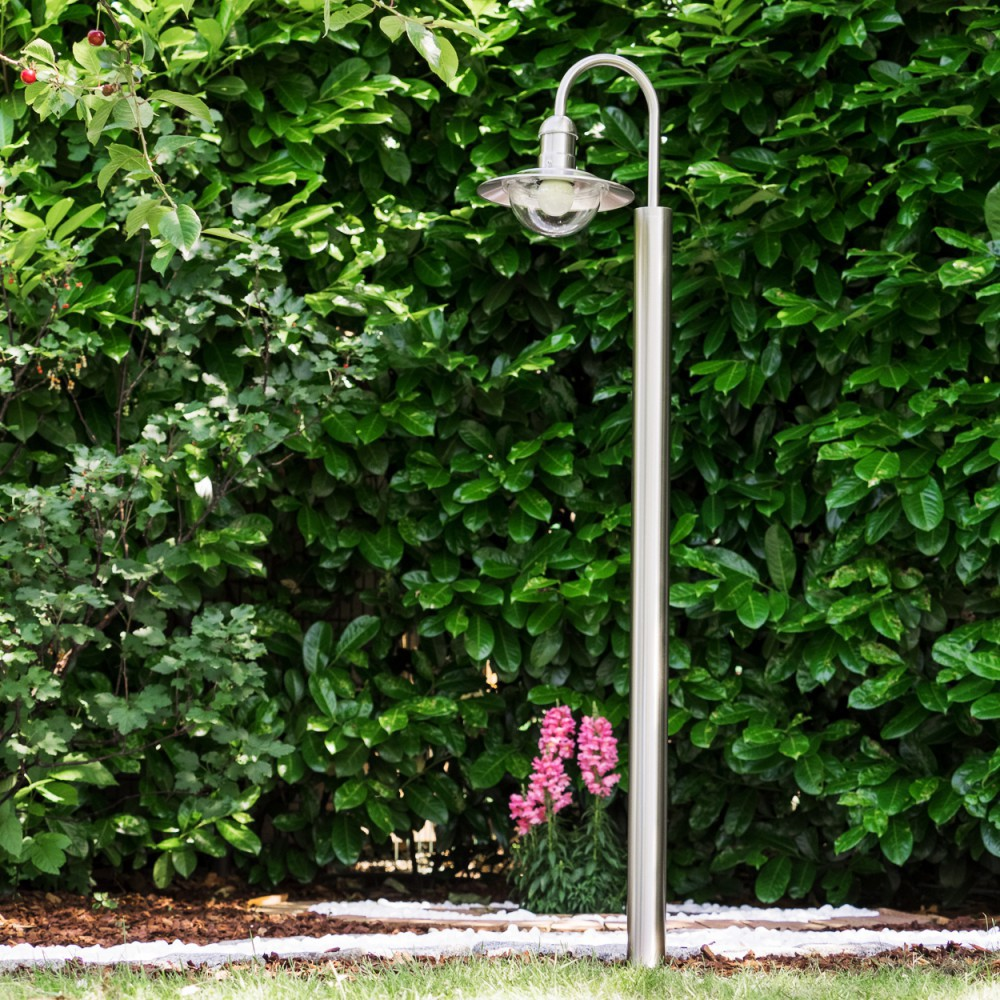 Ein schlanker, silberner Laternenpfahl steht vor einer dichten, grünen Hecke. Daneben wachsen rosa Blumen.