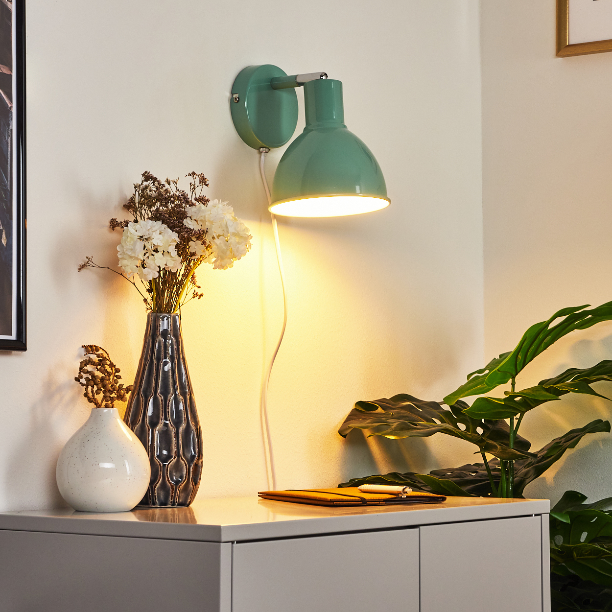 Ein grüner Wandleuchter beleuchtet Blumen in Vasen auf einem grauen Schrank.