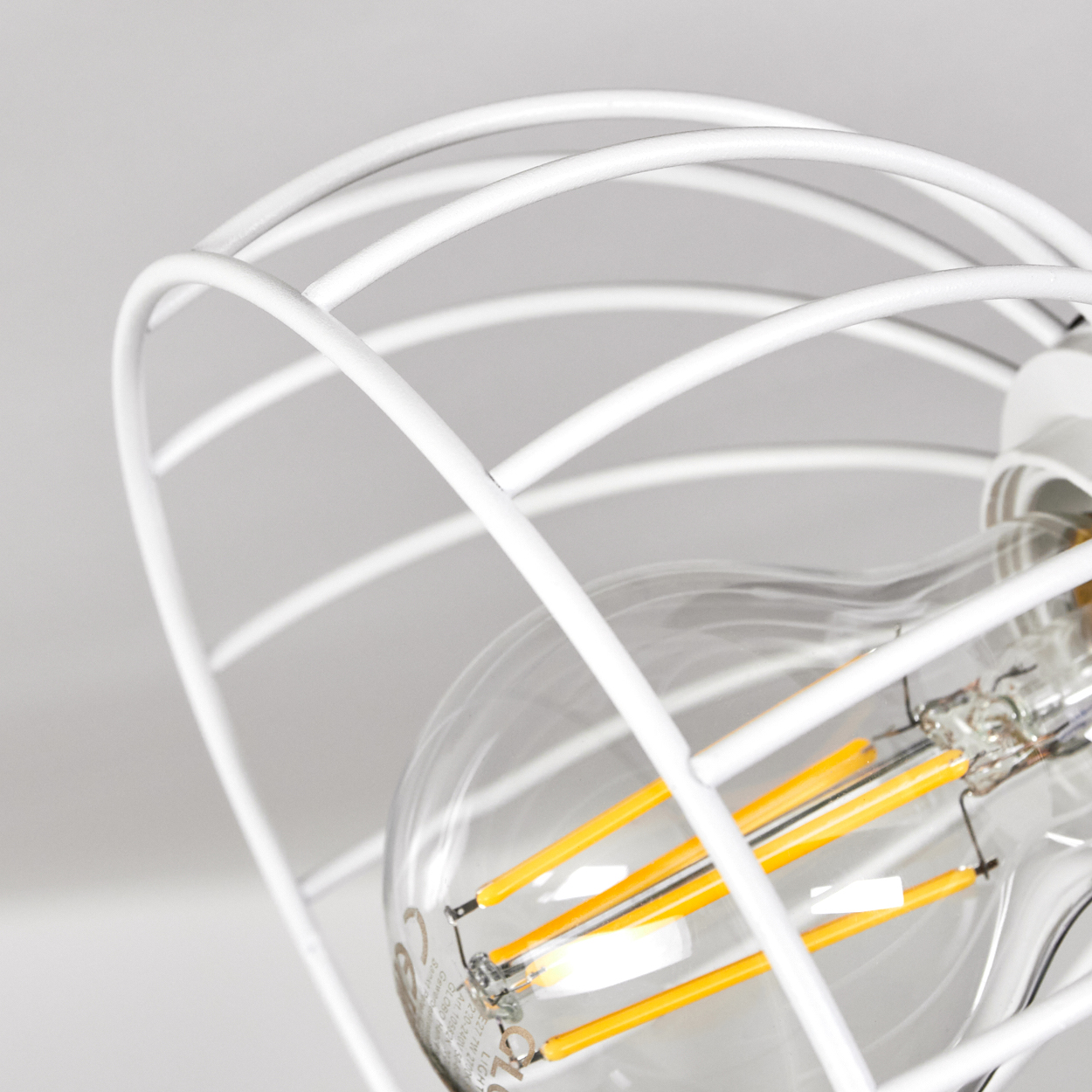 Eine weiße Drahtlampe schützt eine Glühbirne mit sichtbaren gelben Filamenten.