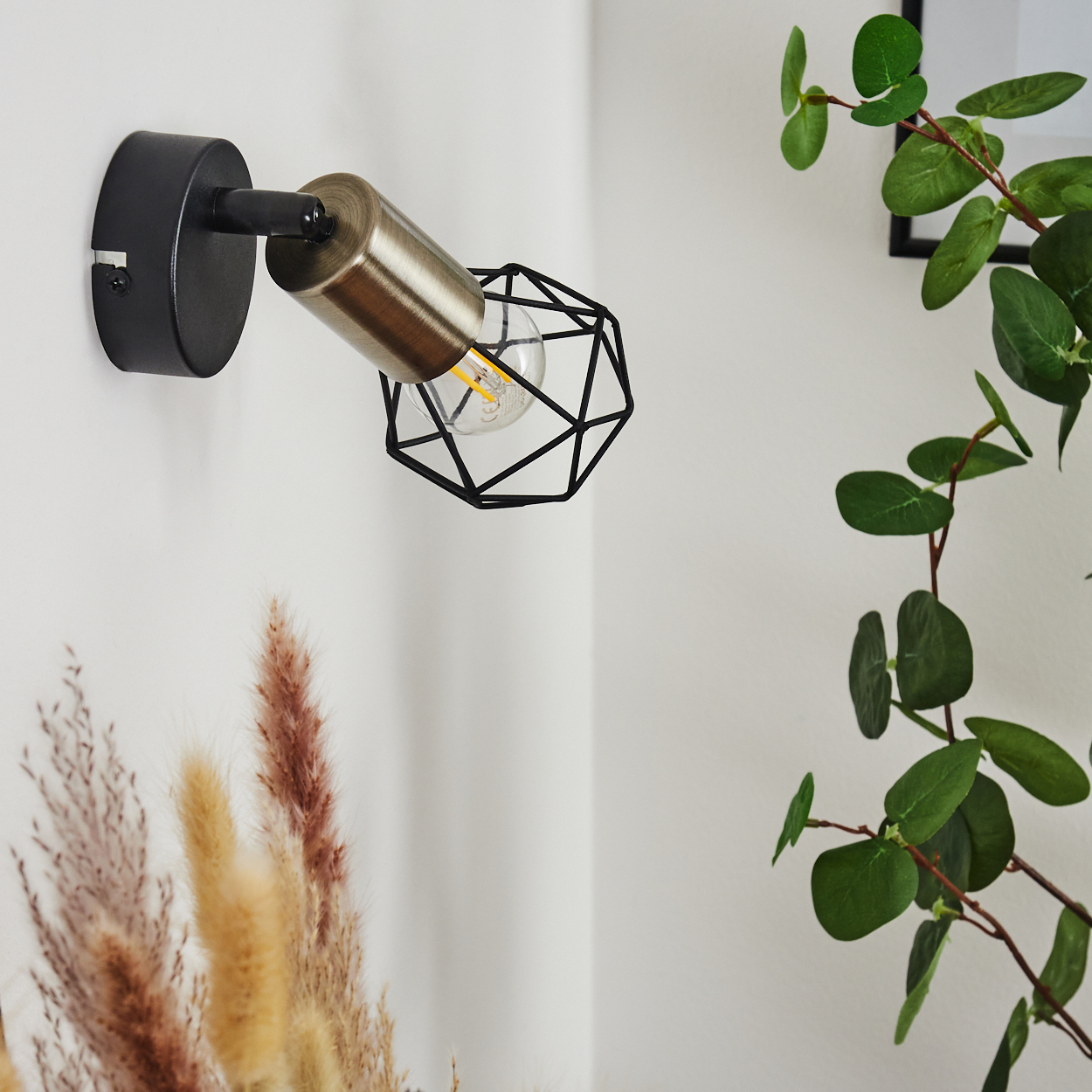 Eine schwarze Wandleuchte mit Metallkäfig und einem sichtbaren Leuchtmittel an einer weißen Wand.