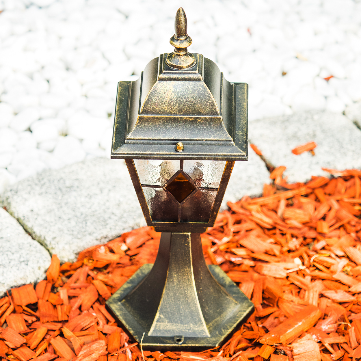 Eine bronzefarbene Gartenlaterne steht auf rotem Rindenmulch neben weißen Steinen.