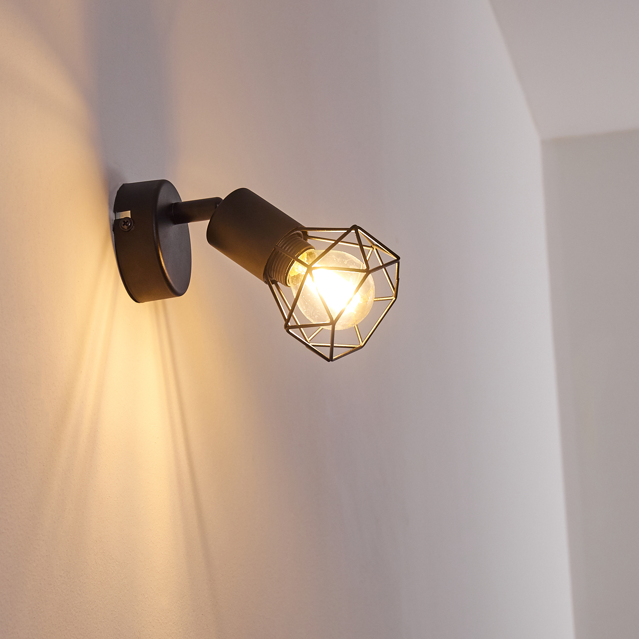 Ein schwarzer Wandstrahler mit Glühbirne und Gitterkäfig beleuchtet eine weiße Wand.