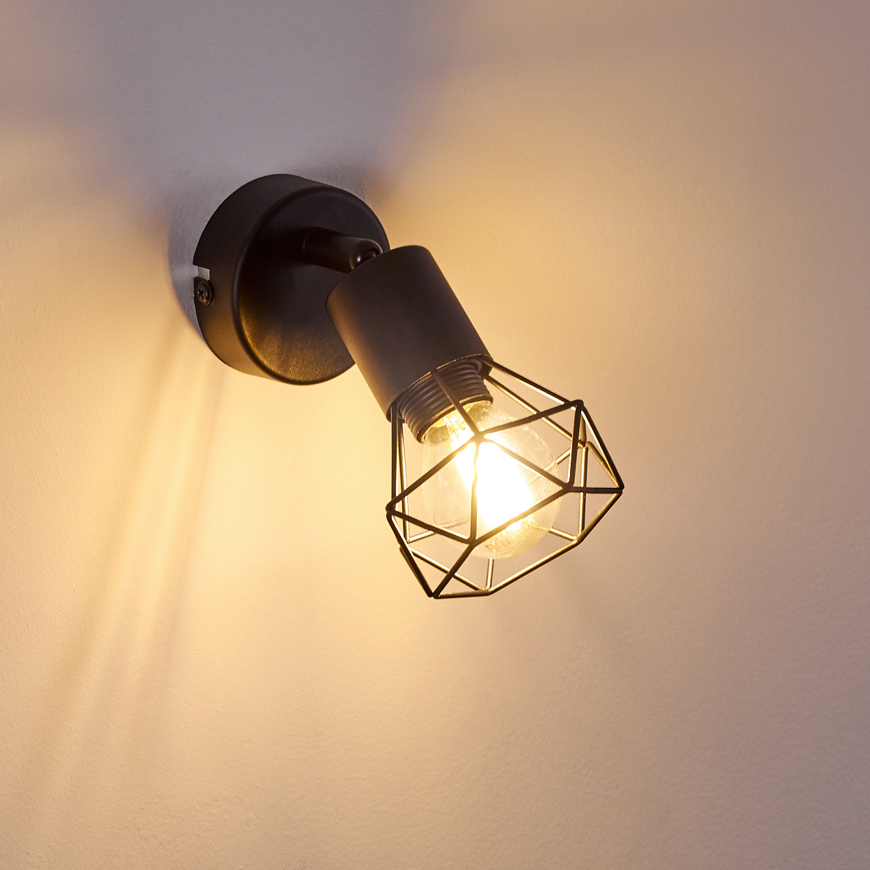 Schwarze Wandleuchte mit Käfig-Lampenschirm und Glühbirne, die Licht abstrahlt.