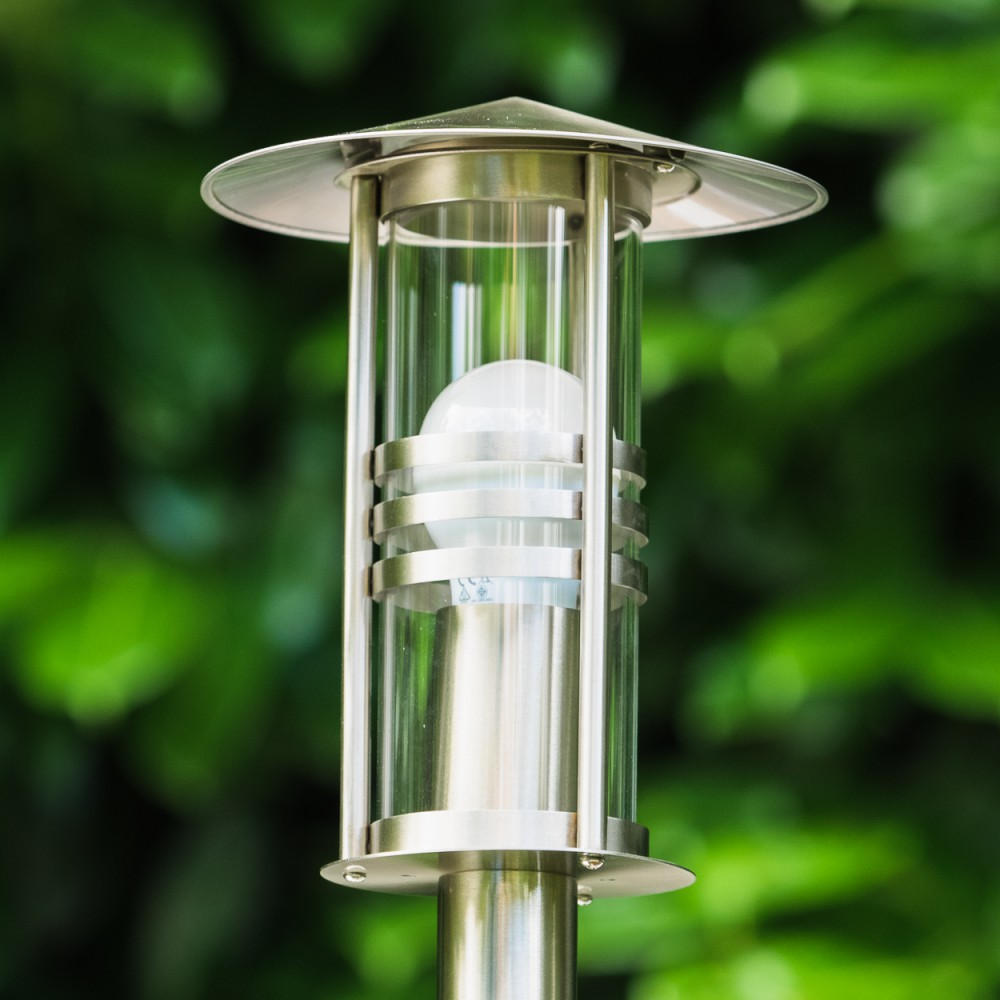 Eine moderne Edelstahl-Gartenleuchte mit Glaszylinder und Glühbirne im Freien.