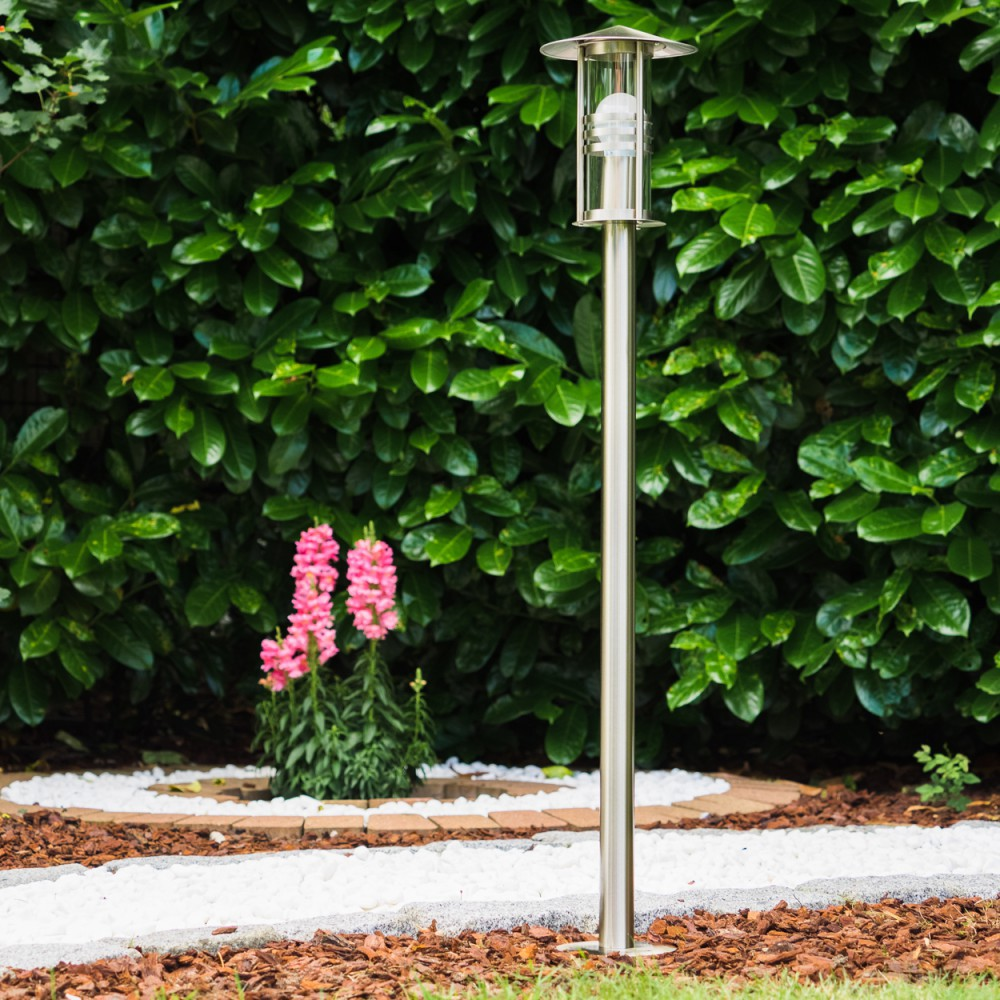 Ein moderner Edelstahl-Gartenlichtmast steht vor einem dichten grünen Busch.
