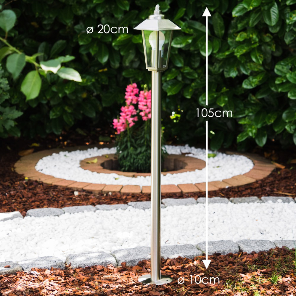 Eine schlanke, silberne Stehlampe steht im Garten vor grünen Büschen.