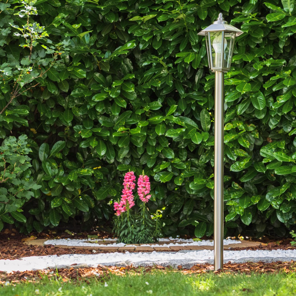 Ein Gartenweg mit weißem Kies und einer Lampe vor einer grünen Hecke mit Blumen.