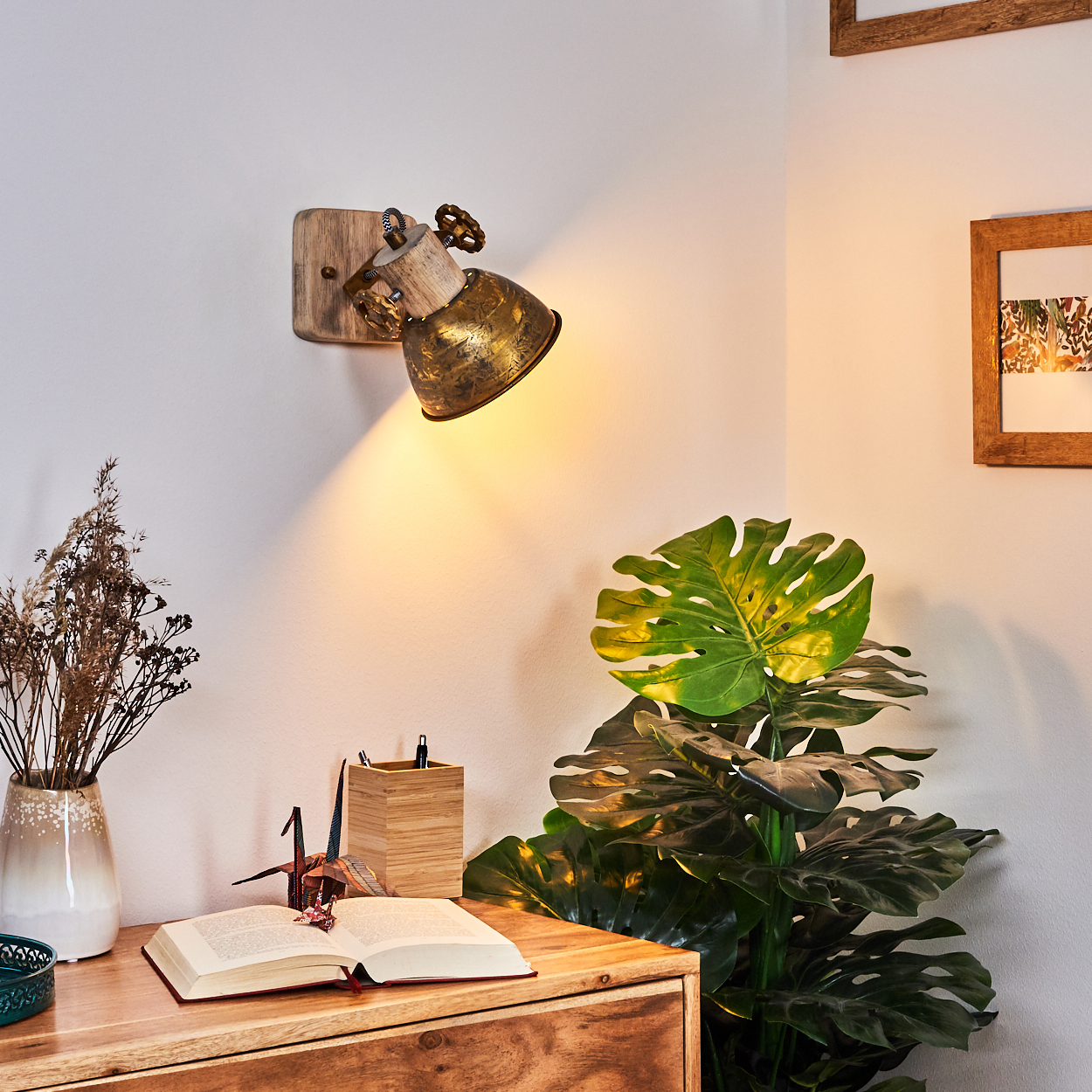 Ein Wandstrahler beleuchtet eine Holzkommode mit Buch, Papierkranich, Vase und Pflanze.
