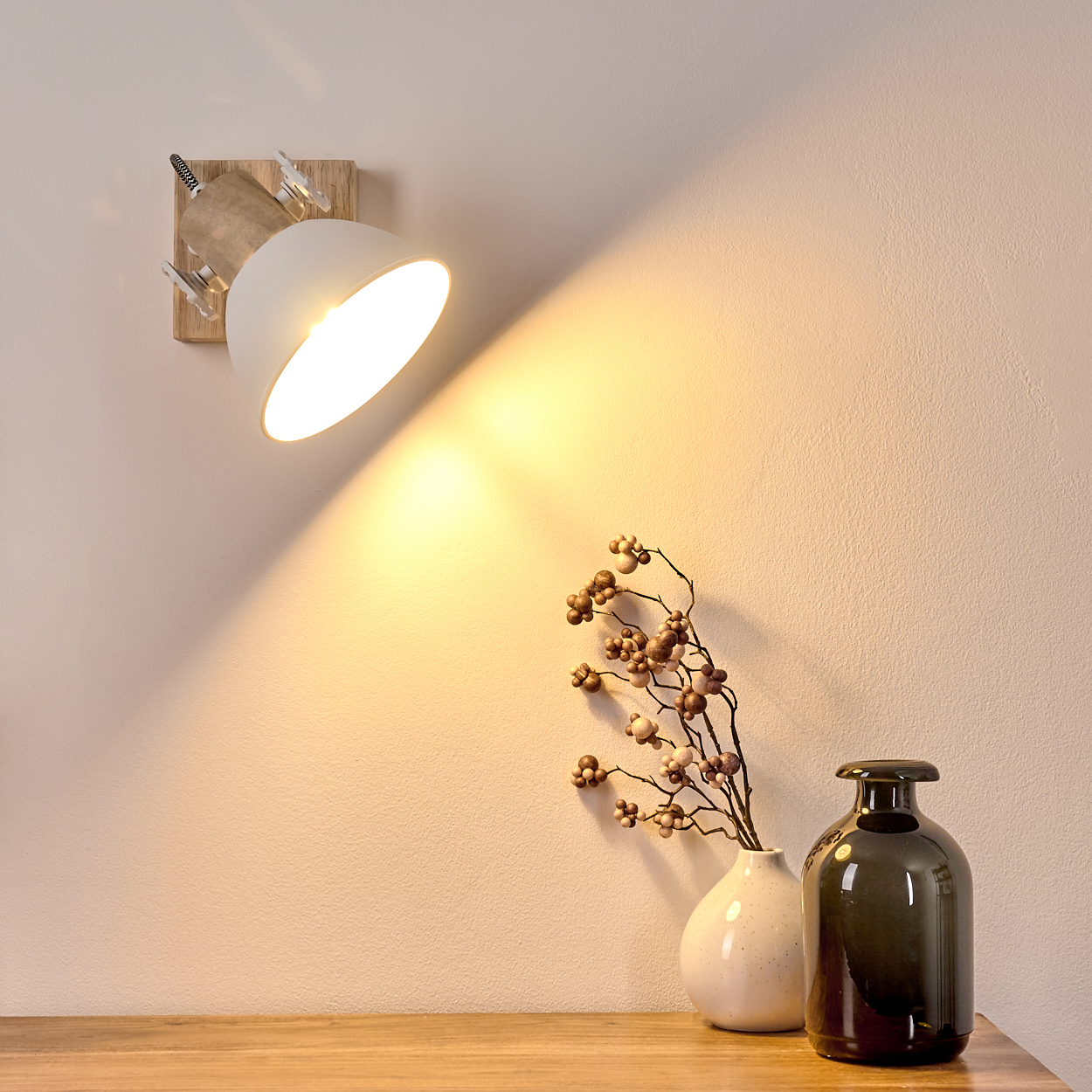 Eine Wandlampe mit Holzsockel beleuchtet eine Wand. Daneben stehen zwei Vasen mit Zweigen.