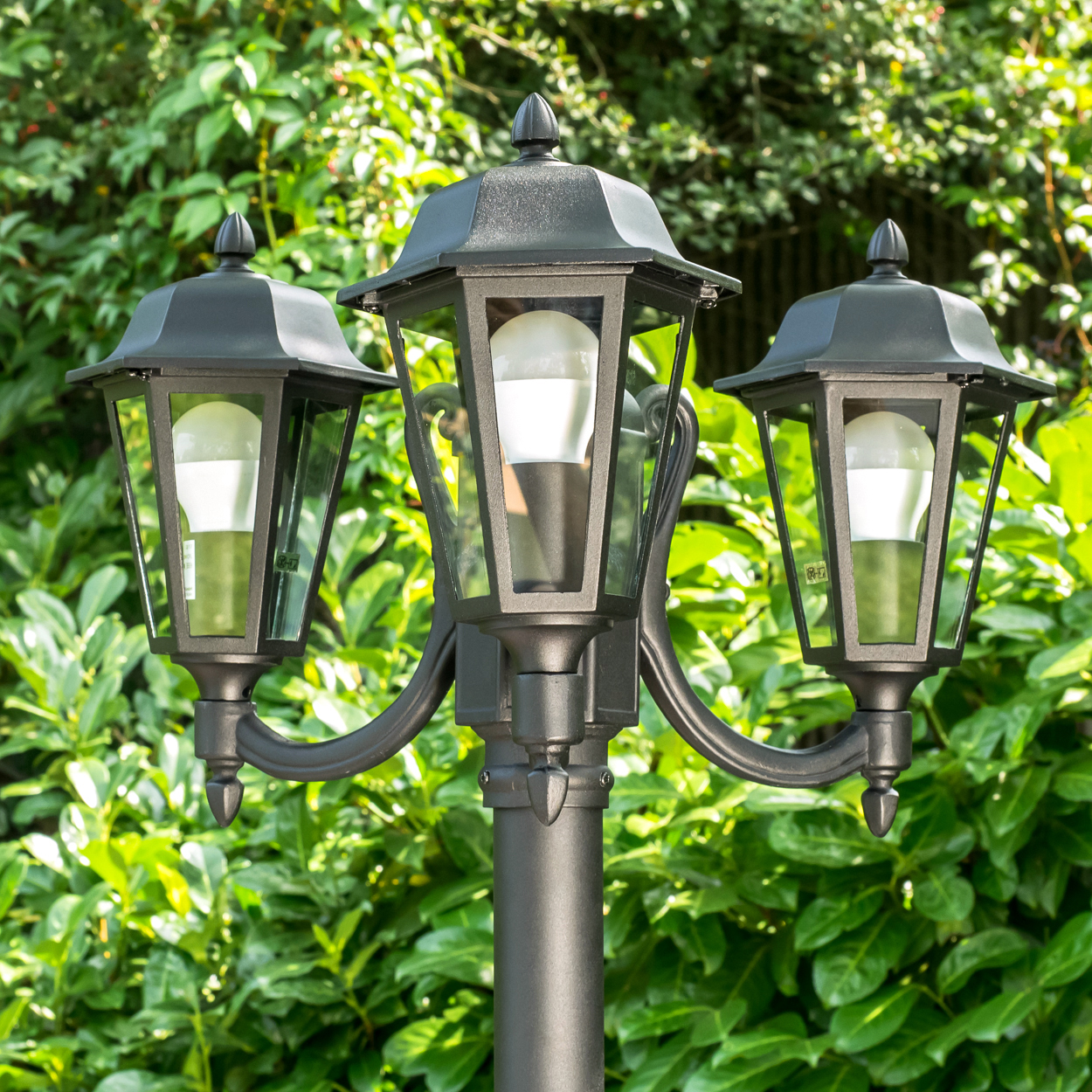 Schwarzer Laternenpfahl mit drei Laternen und LED-Lampen vor grünem Laub.