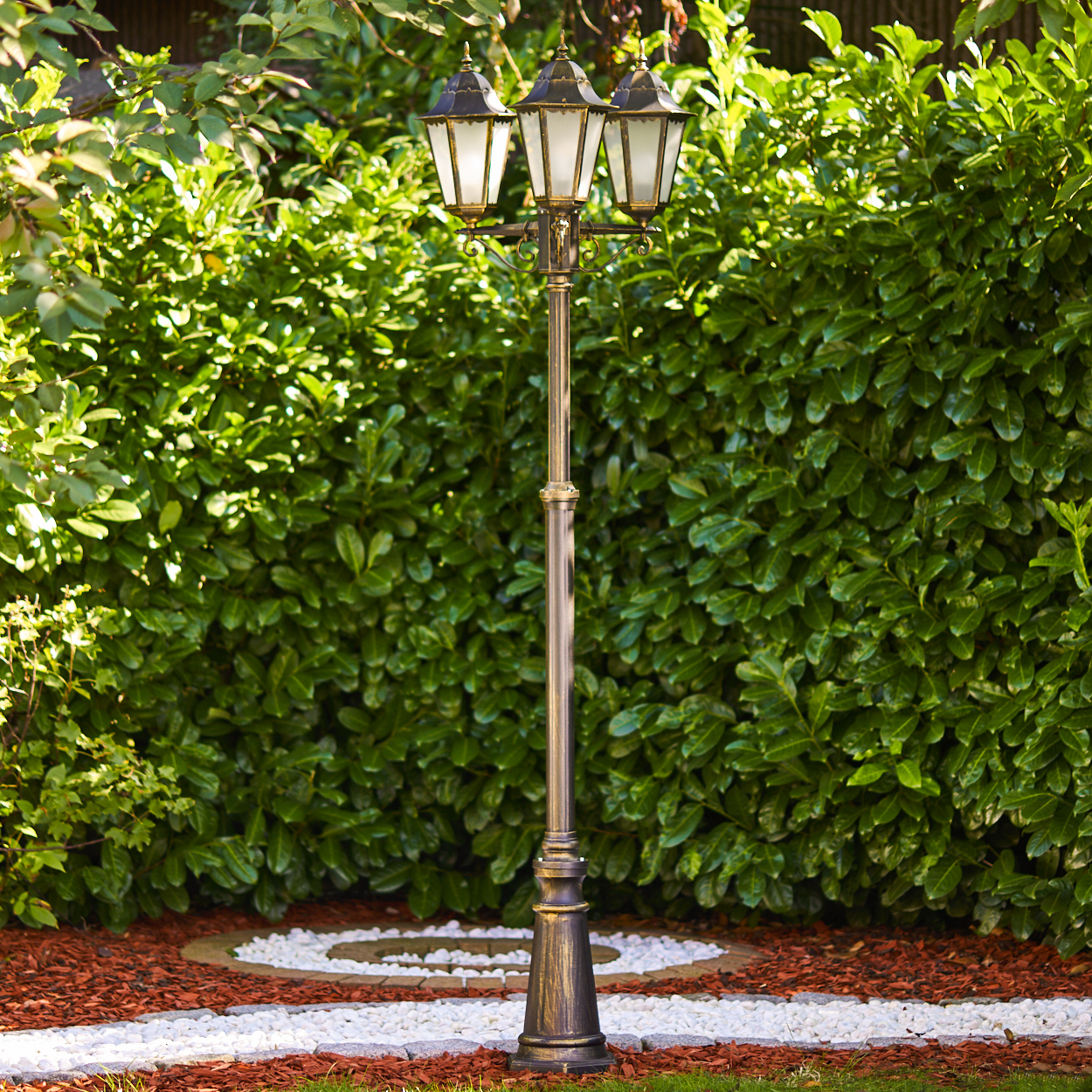 Ein dreiflammiger Gartenlaternenmast aus Metall steht vor einer dichten Hecke.