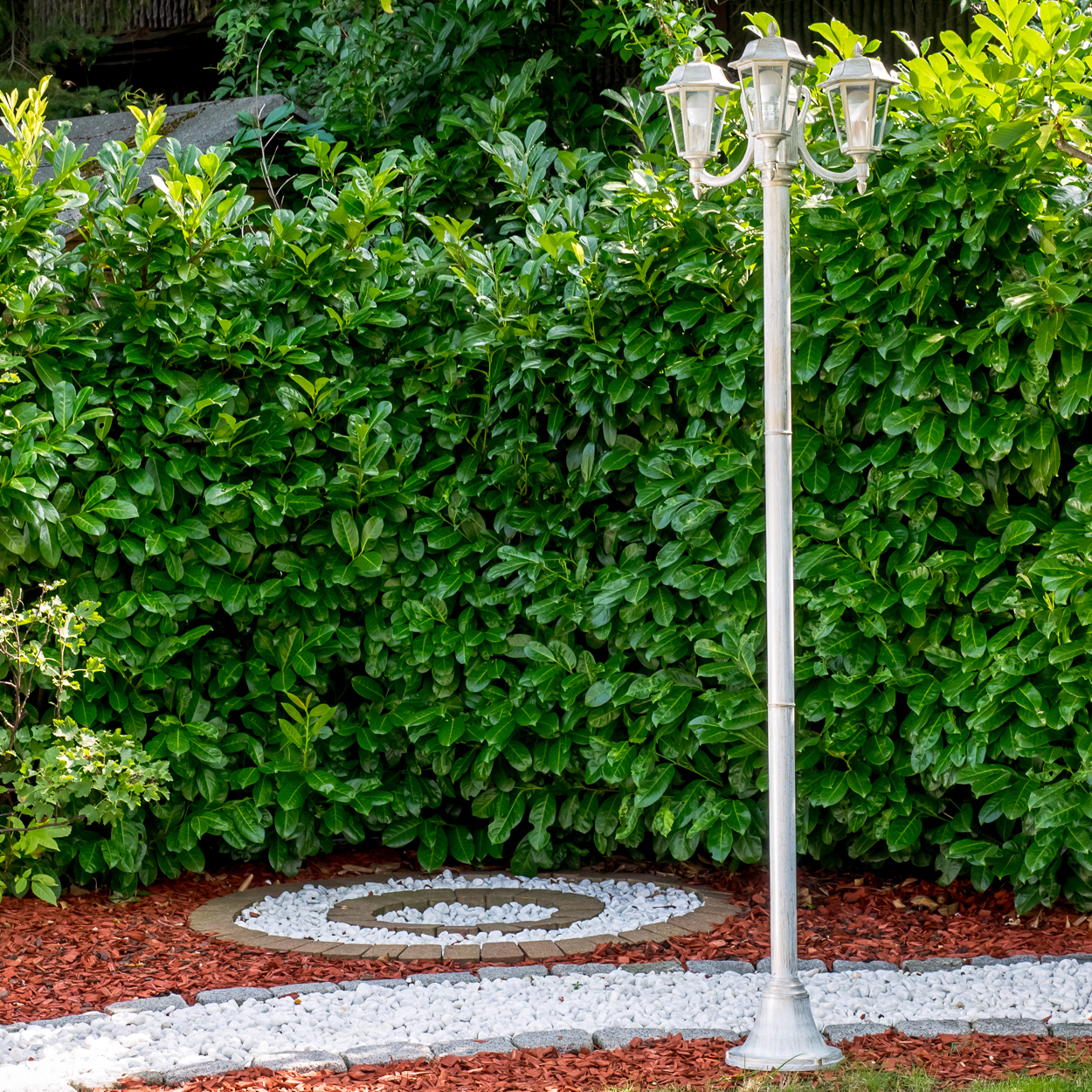 Ein weißer Laternenpfahl steht vor einer dichten grünen Hecke. Dekorative Steine schmücken den Boden.