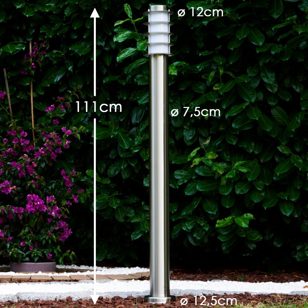 Ein schlanker Edelstahl-Gartenpfosten mit weißem Lichtkopf steht vor grünen Büschen.