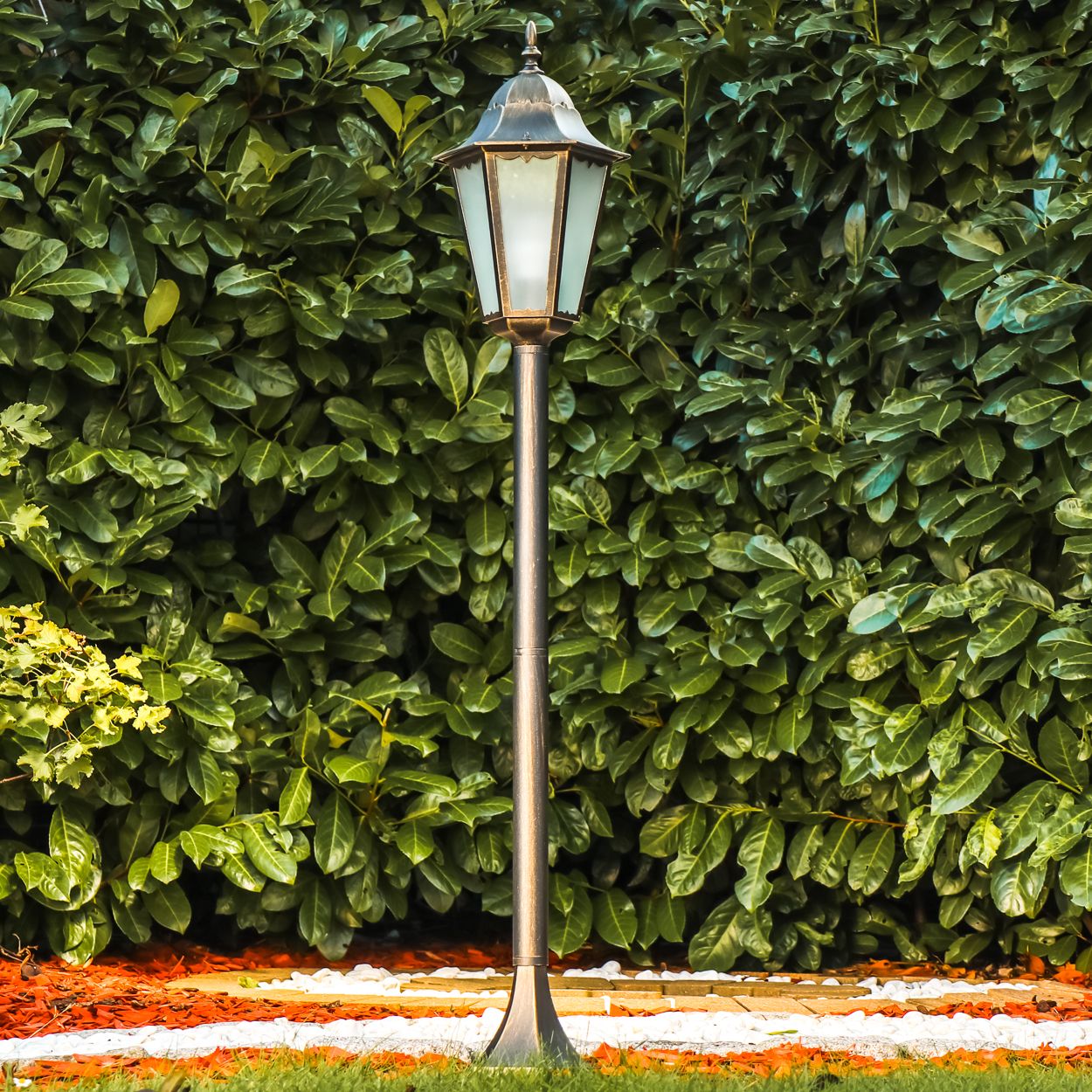 Ein Laternenpfahl steht vor einer dichten grünen Hecke. Im Vordergrund liegen orangefarbene Blätter.