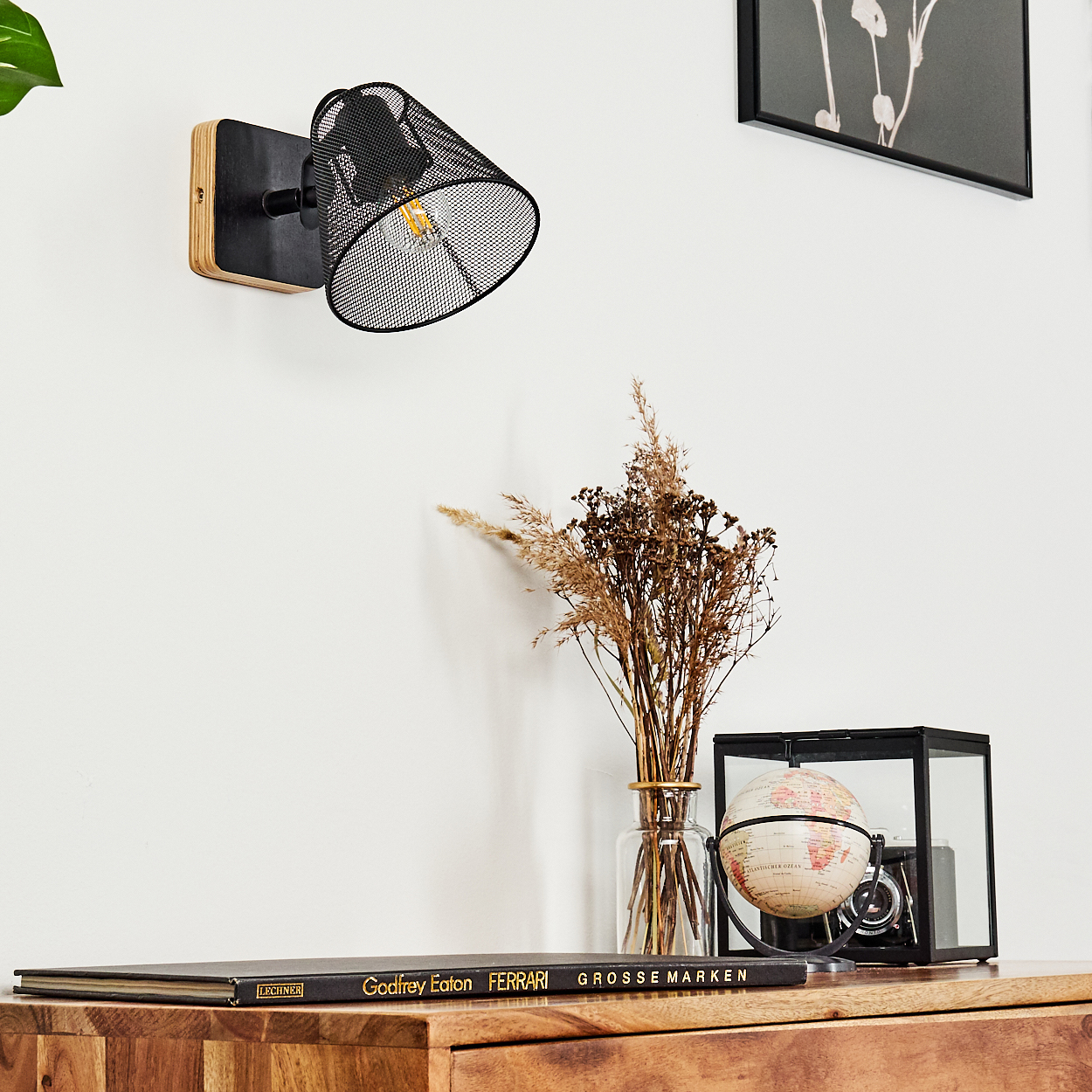 Ein schwarzer Wandleuchter aus Netzstoff beleuchtet eine Holzkommode mit Deko: Trockenblumen, Globus, Kamera, Buch.