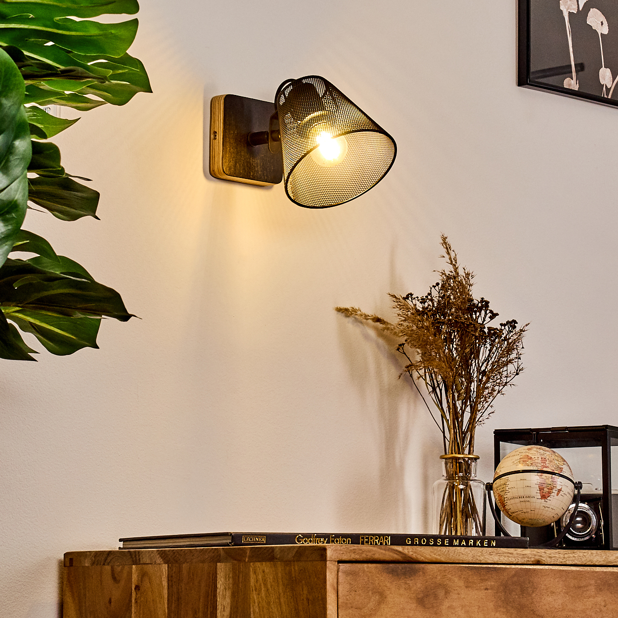 Ein schwarzer Wandspot mit Gitterschirm beleuchtet eine weiße Wand neben Pflanzen.