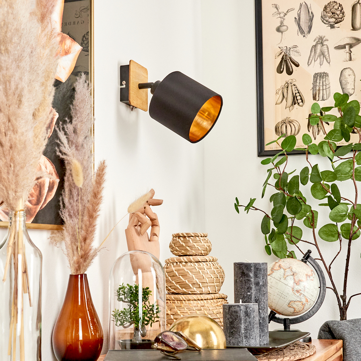 Eine schwarze Wandleuchte mit goldenem Inneren hängt über Dekoartikeln wie Pflanzen und Kerzen.