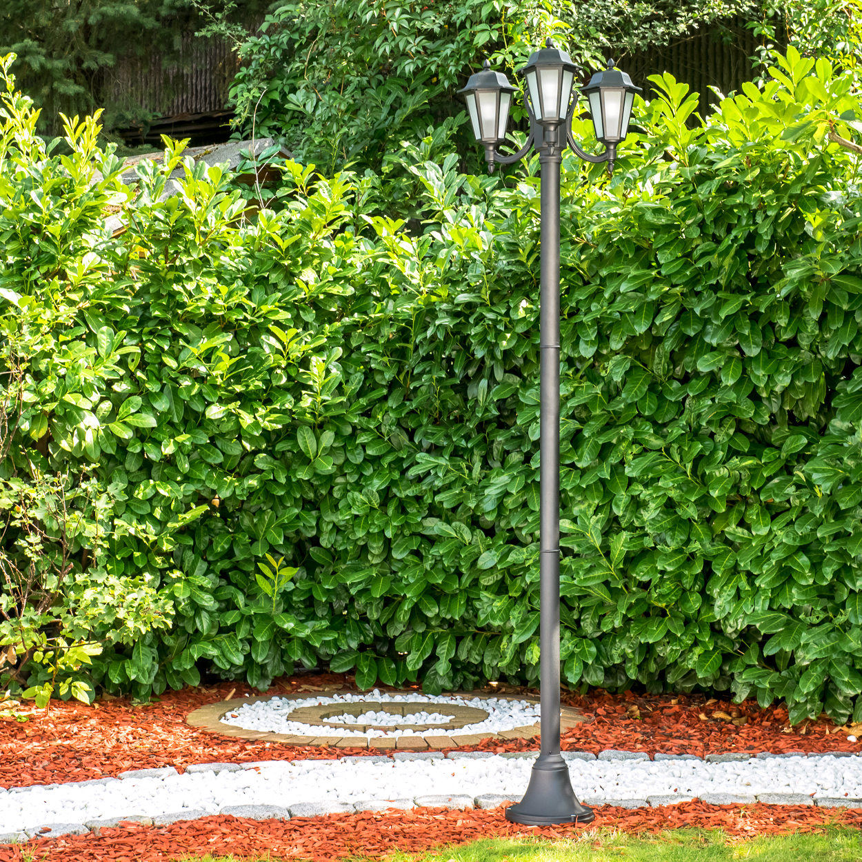 Ein dunkler Laternenpfahl steht vor einer dichten grünen Hecke. Der Boden ist mit Rindenmulch bedeckt.