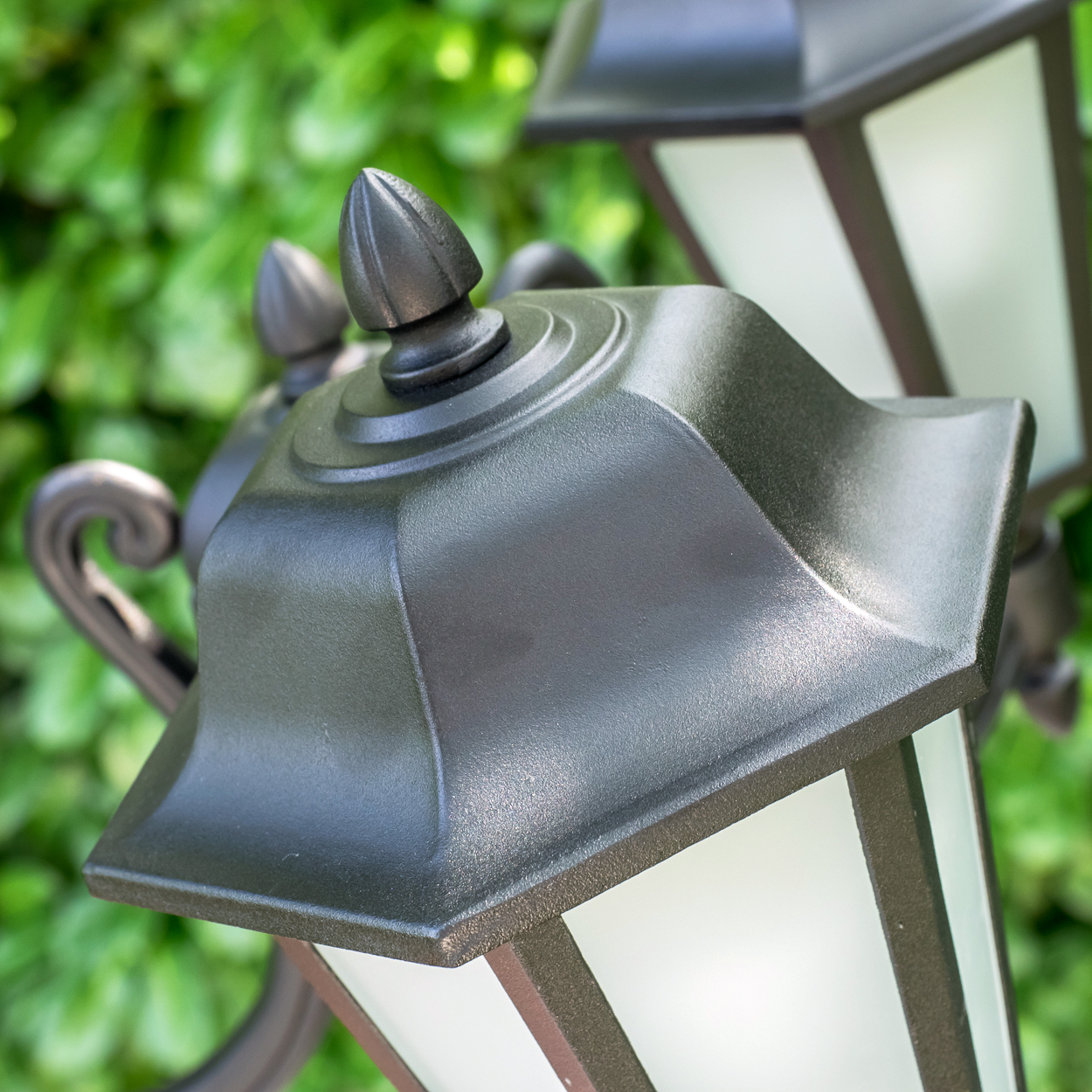 Dunkle Außenlampe mit spitzem Dekor über einem Milchglas-Panel, vor grünem Laub.