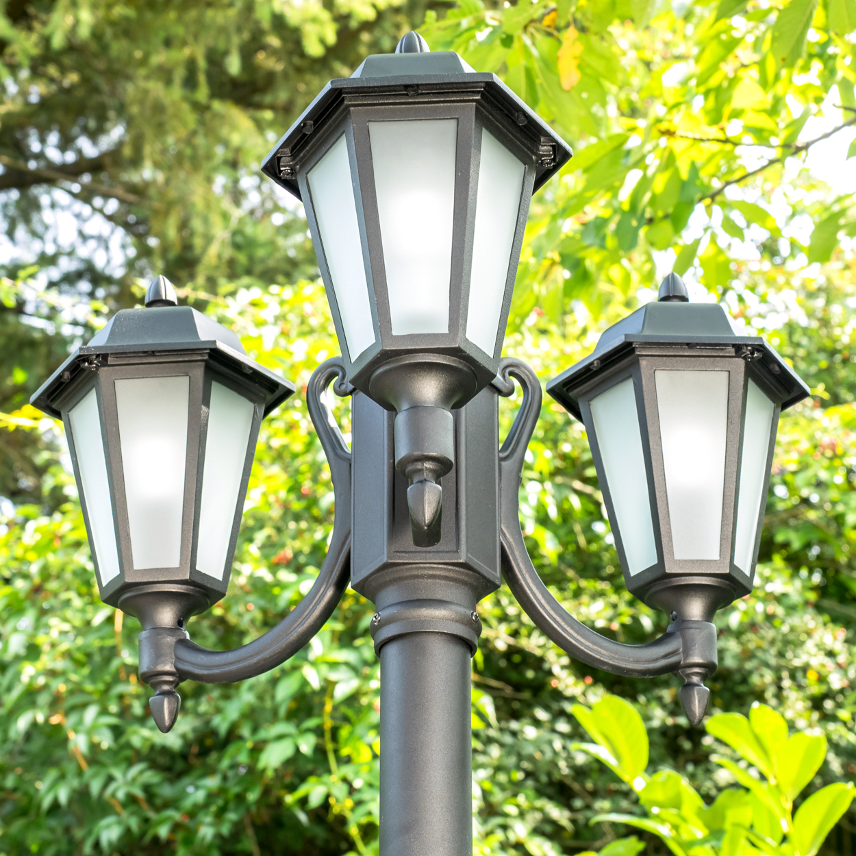 Ein schwarzer Laternenmast mit drei Lichtern vor grünem Laub.