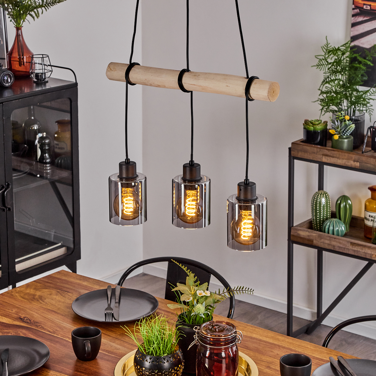 Hängeleuchte mit Holzstange und drei Zylindern über einem Holztisch.