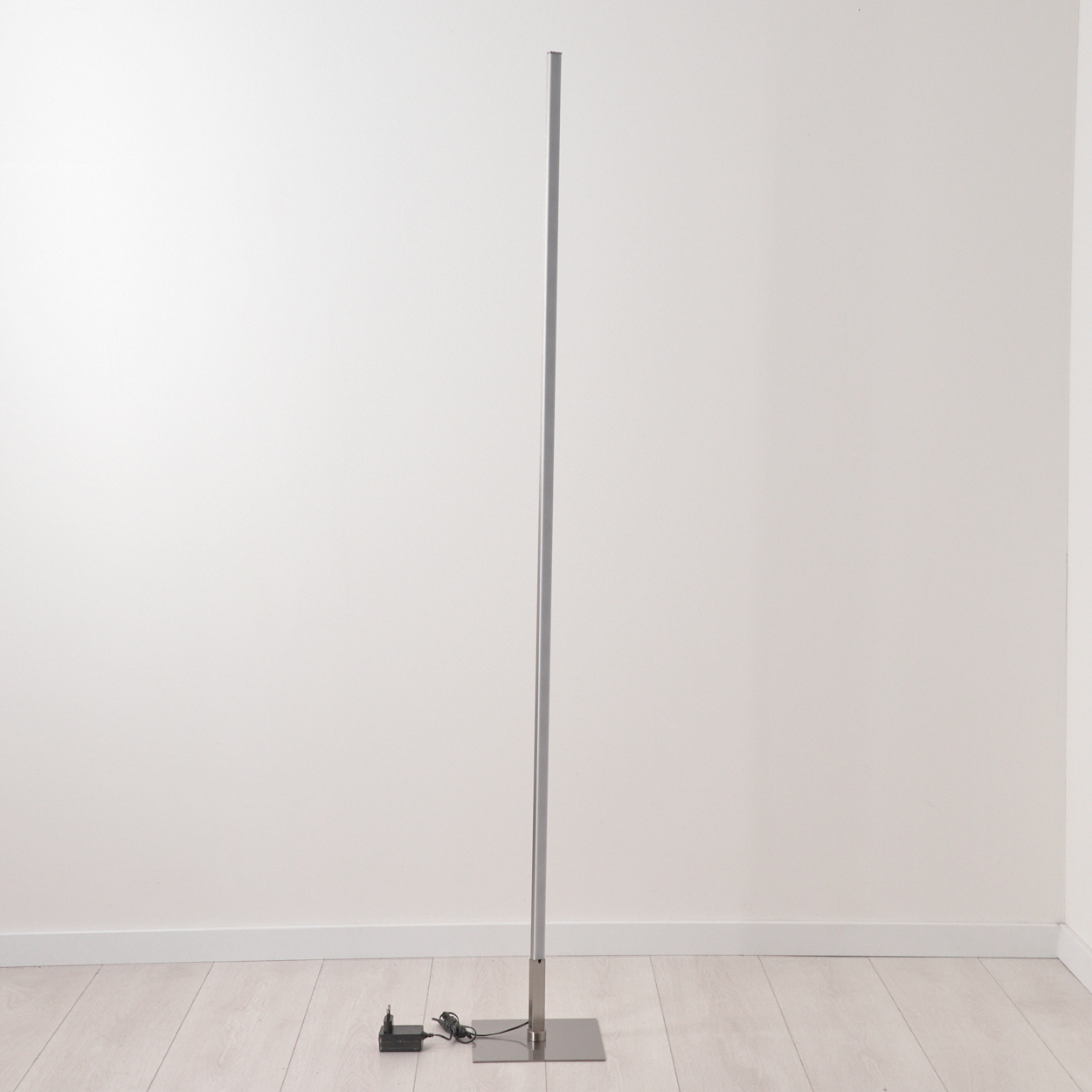 Ein schlanker, silberner Stehleuchter mit rechteckiger Basis und Netzteil.