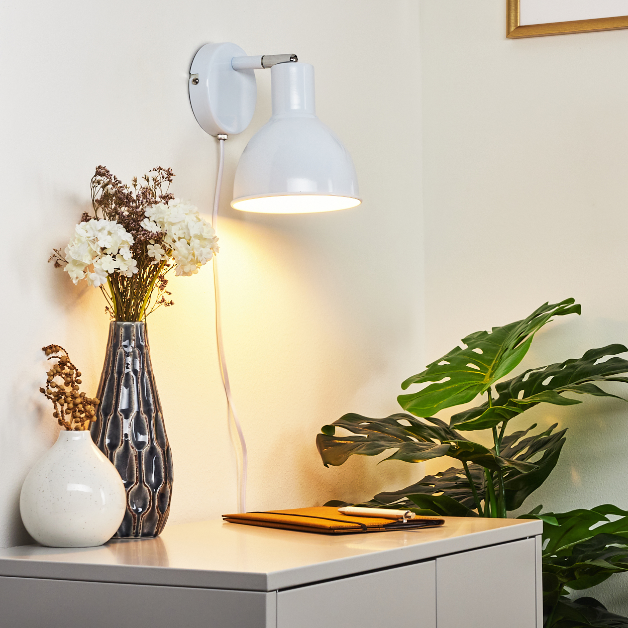 Eine weiße Wandleuchte beleuchtet eine Kommode mit Vasen und Pflanzen.