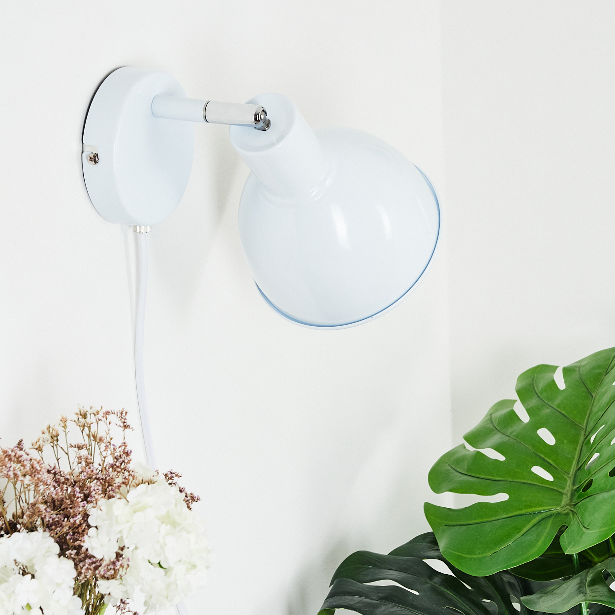 Eine weiße Wandlampe mit Kabel hängt an einer weißen Wand neben Pflanzen und Blumen.