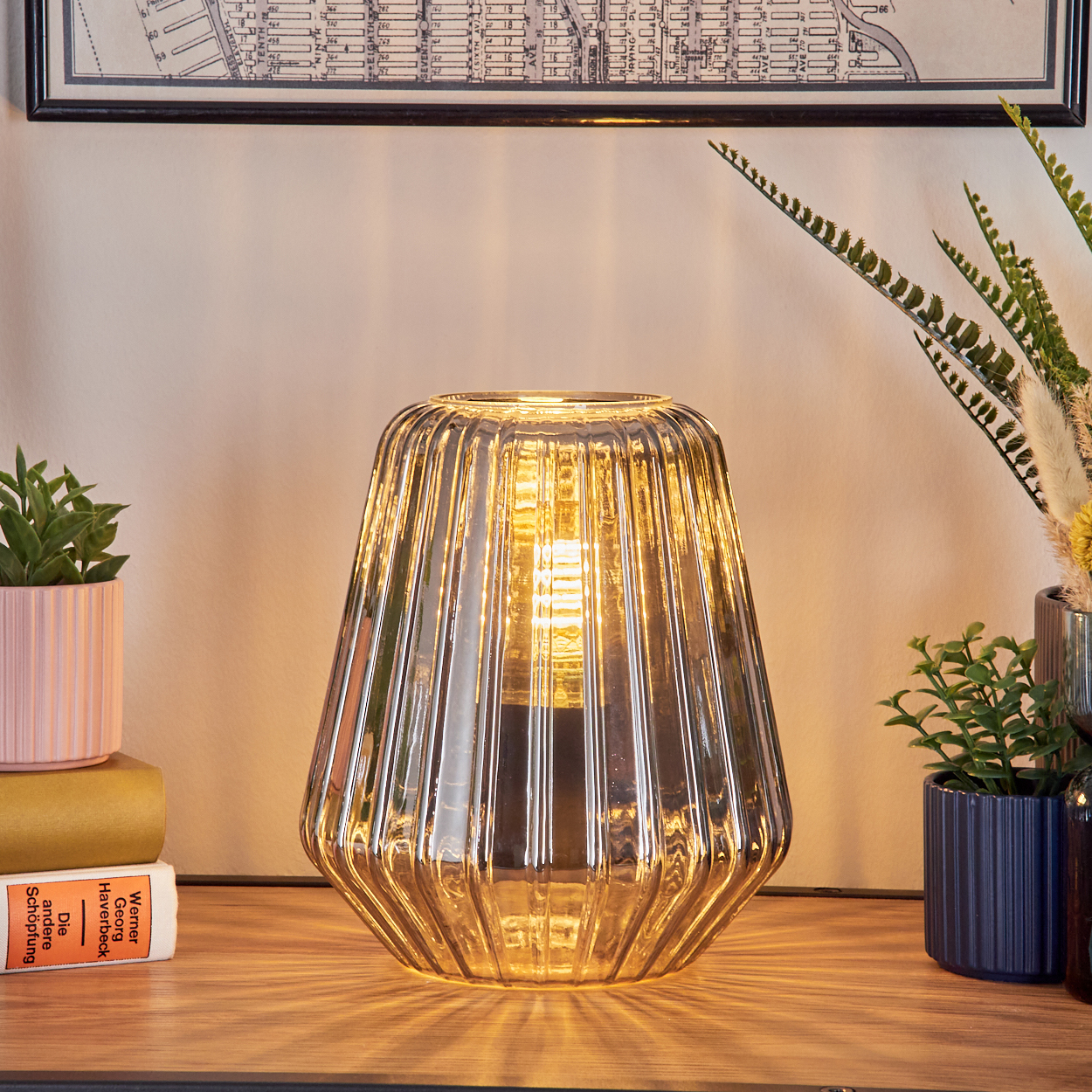 Eine gerippte Glaslampe leuchtet auf einem Holztisch neben Pflanzen und Büchern.
