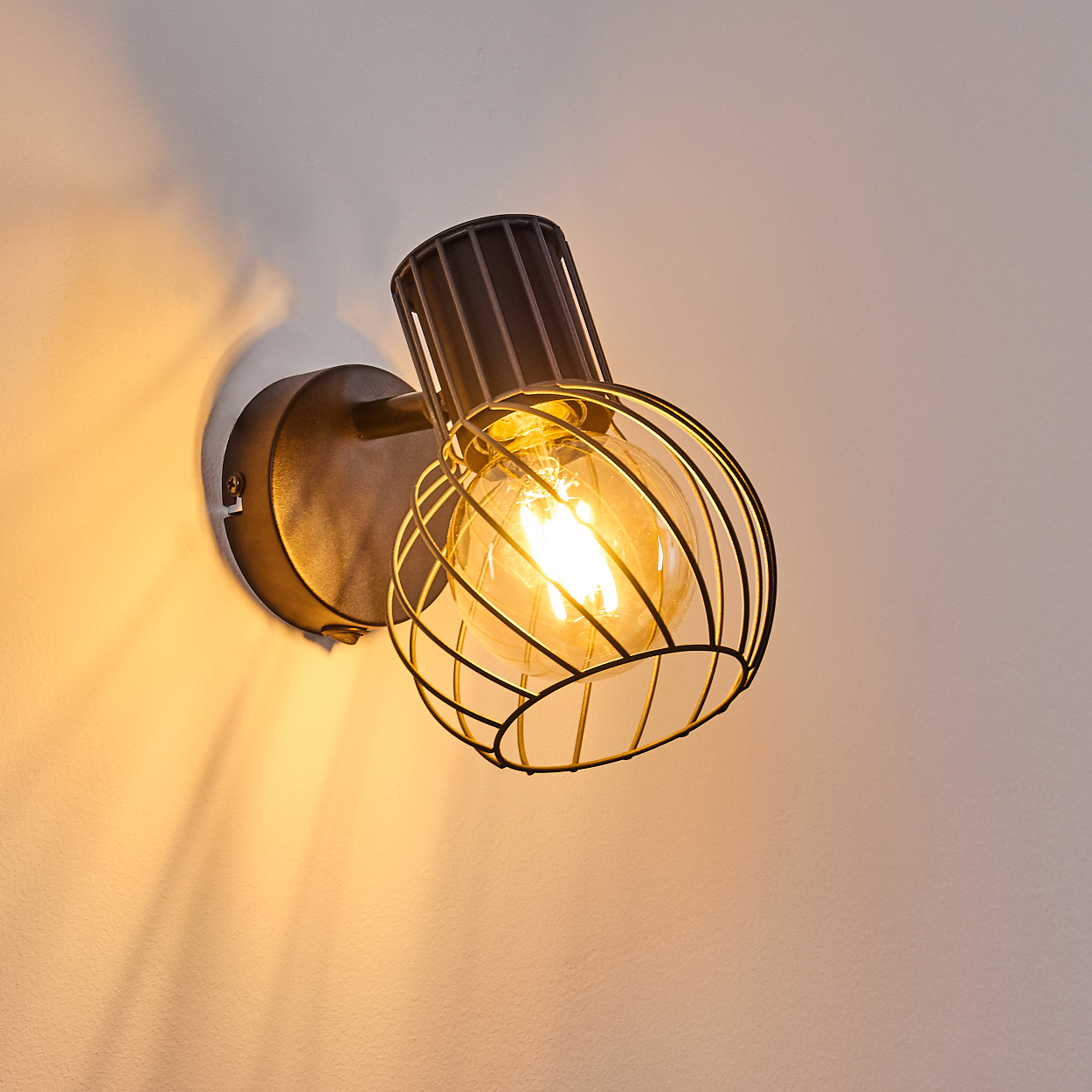Eine dunkle Wandleuchte mit Glühbirne und Drahtkäfig leuchtet an einer weißen Wand.