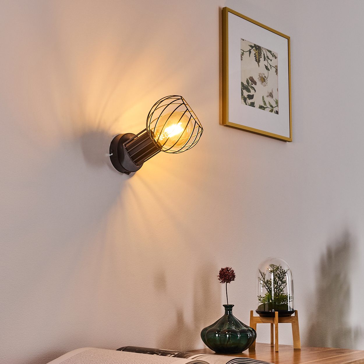 Wandleuchte mit Drahtkäfig über einem Buch, Vase und Glasglocke.