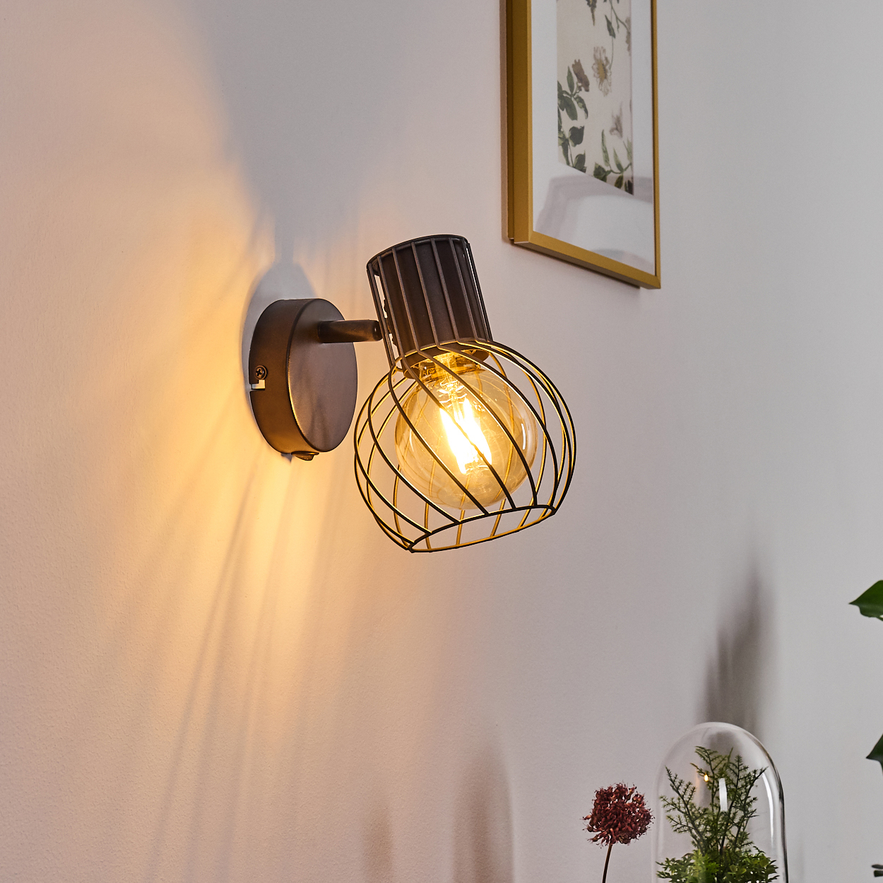 Schwarzer Wandstrahler mit Glühbirne und Drahtkäfig, leuchtet an einer weißen Wand.