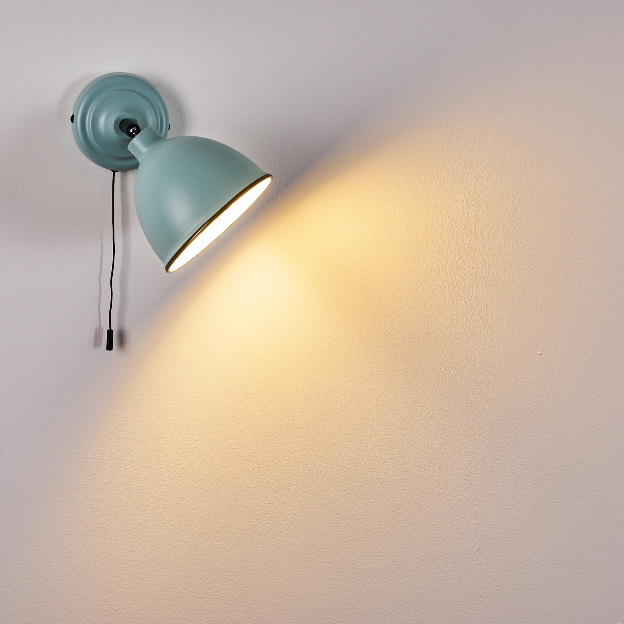 Ein hellblauer Wandspot mit einem schwarzen Zugschalter leuchtet auf eine weiße Wand.