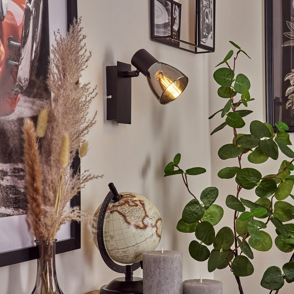 Ein schwarzer Wandspot leuchtet auf einer Wand. Davor ein Globus, zwei Kerzen und Pflanzen.