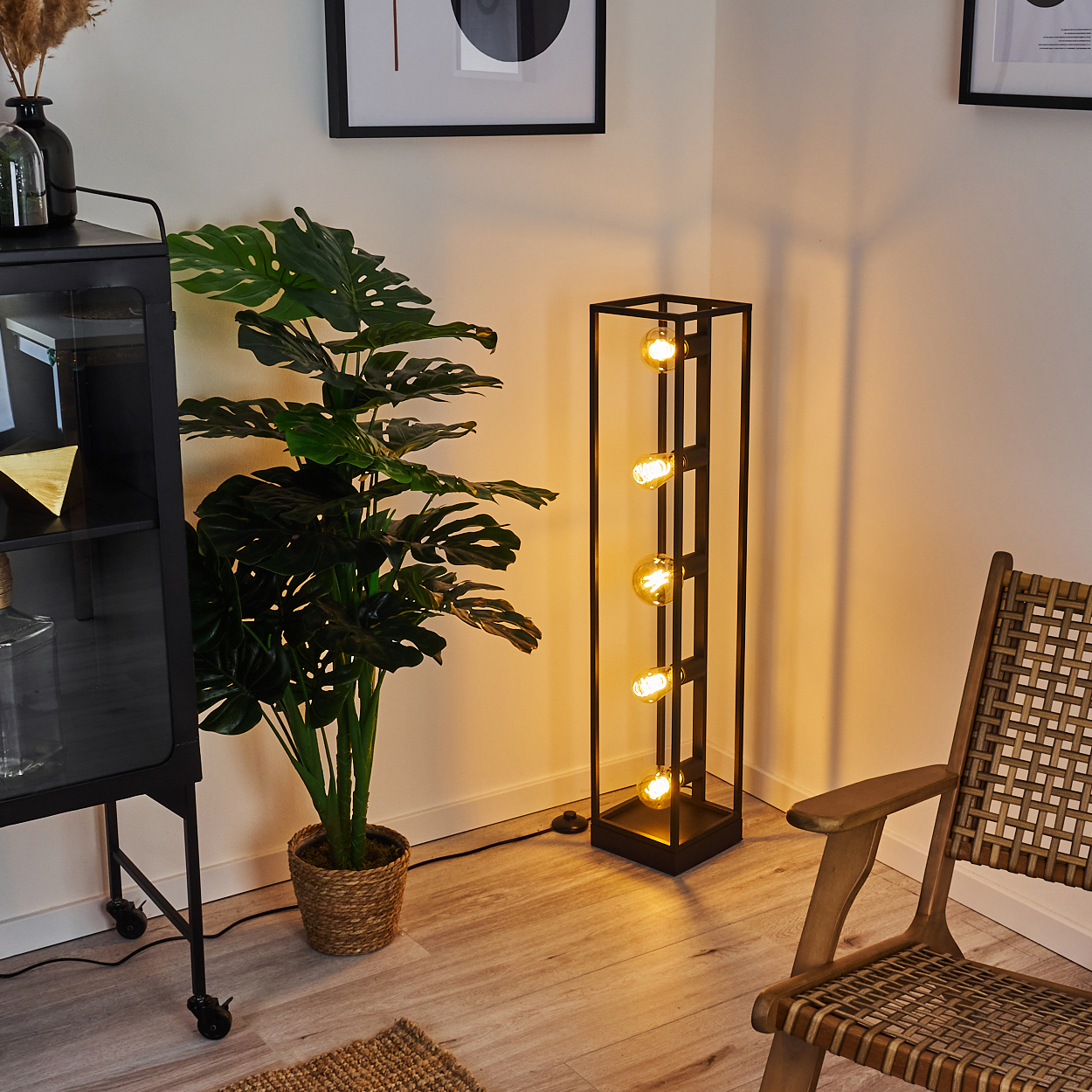 Eine hohe schwarze Stehlampe mit vier Edison-Glühbirnen steht neben einer großen grünen Monstera-Pflanze.