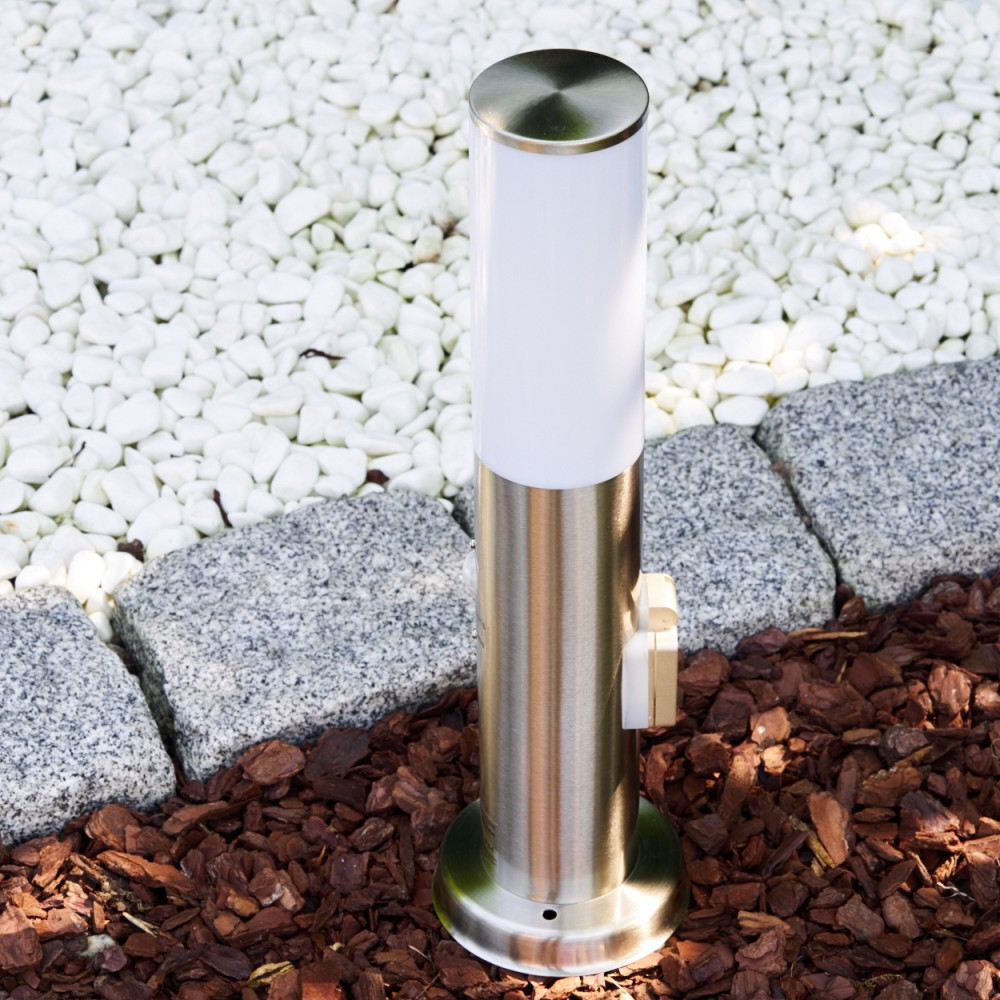 Eine moderne Edelstahl-Gartenleuchte steht auf Rindenmulch neben Steinen.