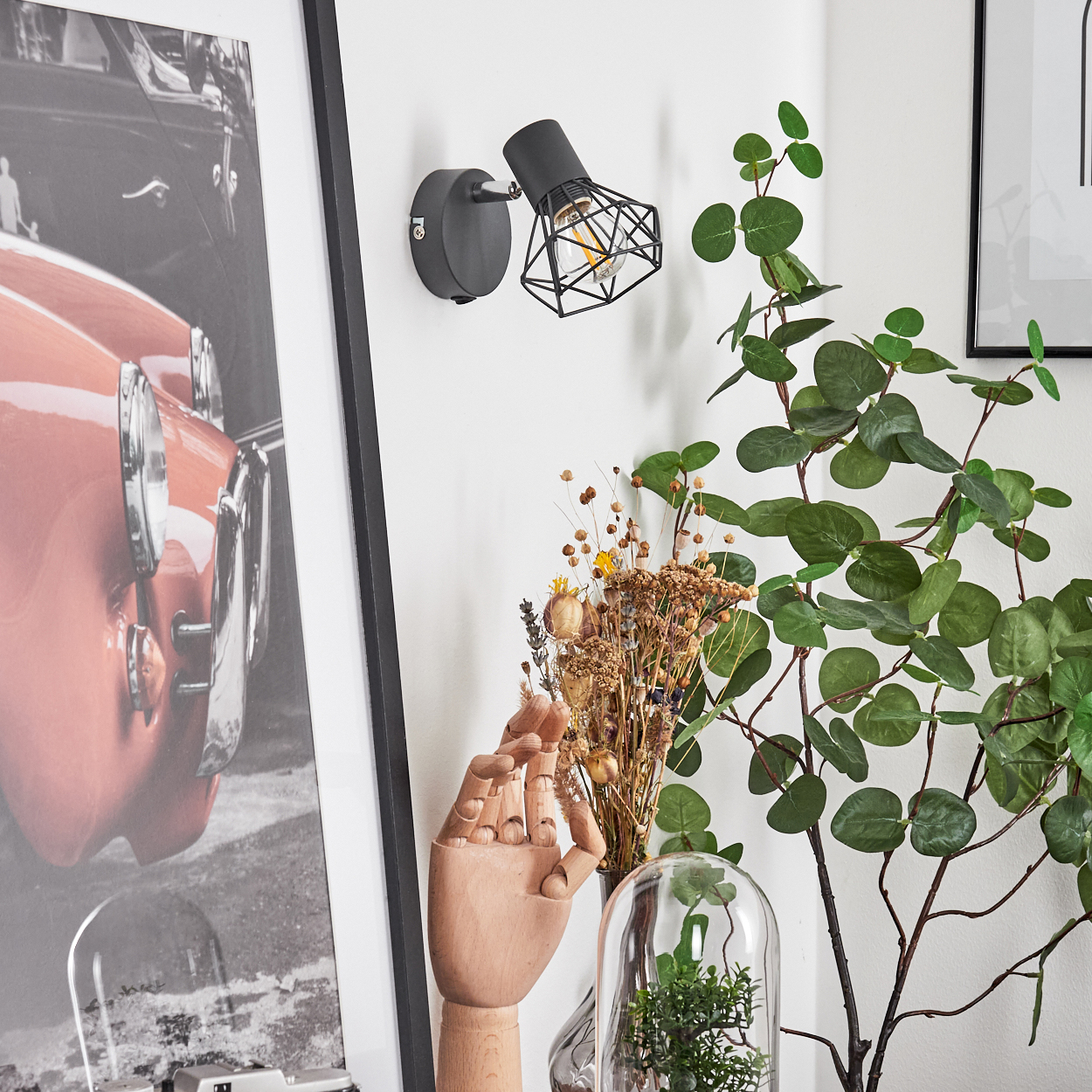 Ein schwarzer Wandspot beleuchtet eine rote Auto-Fotografie, daneben eine Holzhand und Pflanzen.