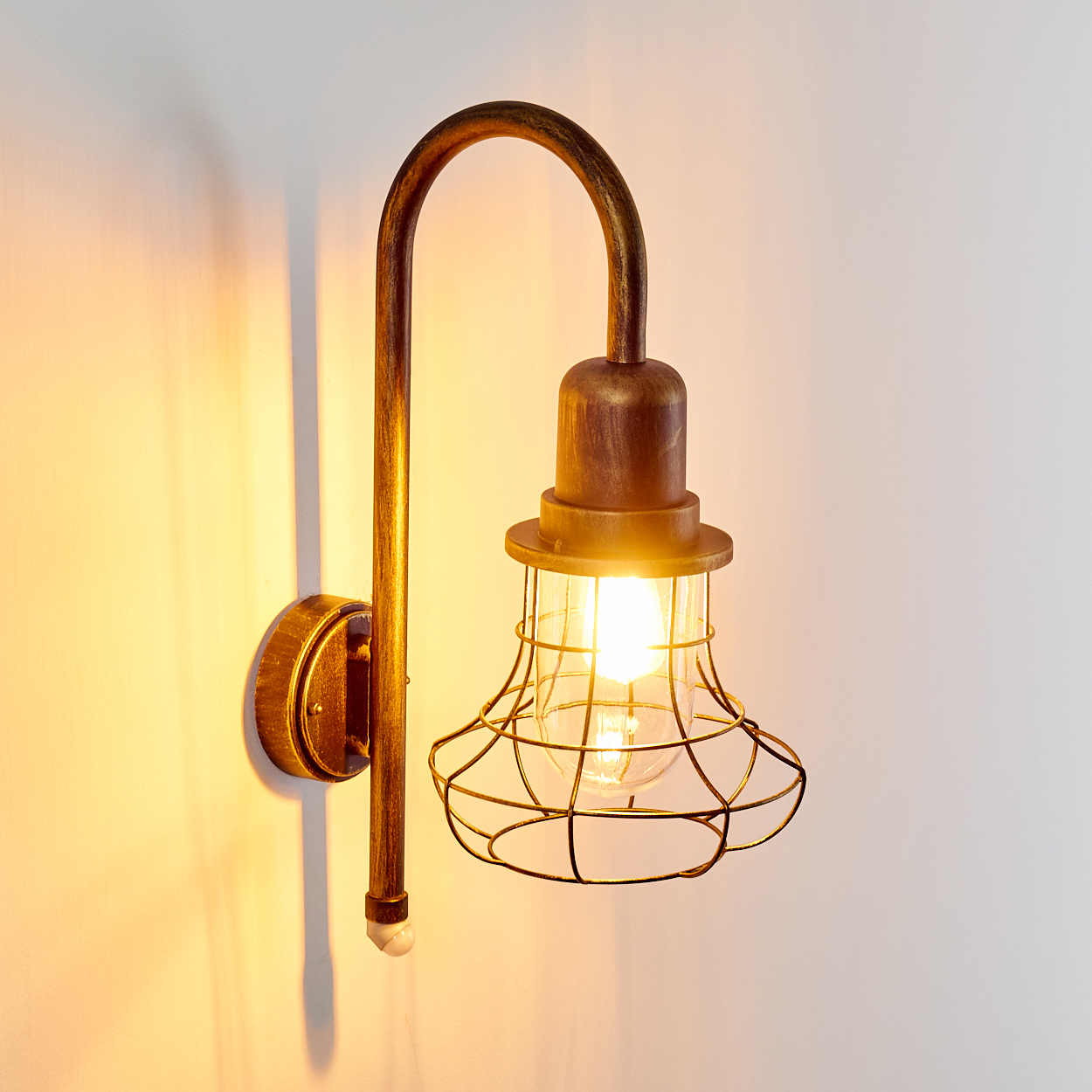 Bronzene Wandleuchte mit Drahtkäfig und Glühbirne an weißer Wand.